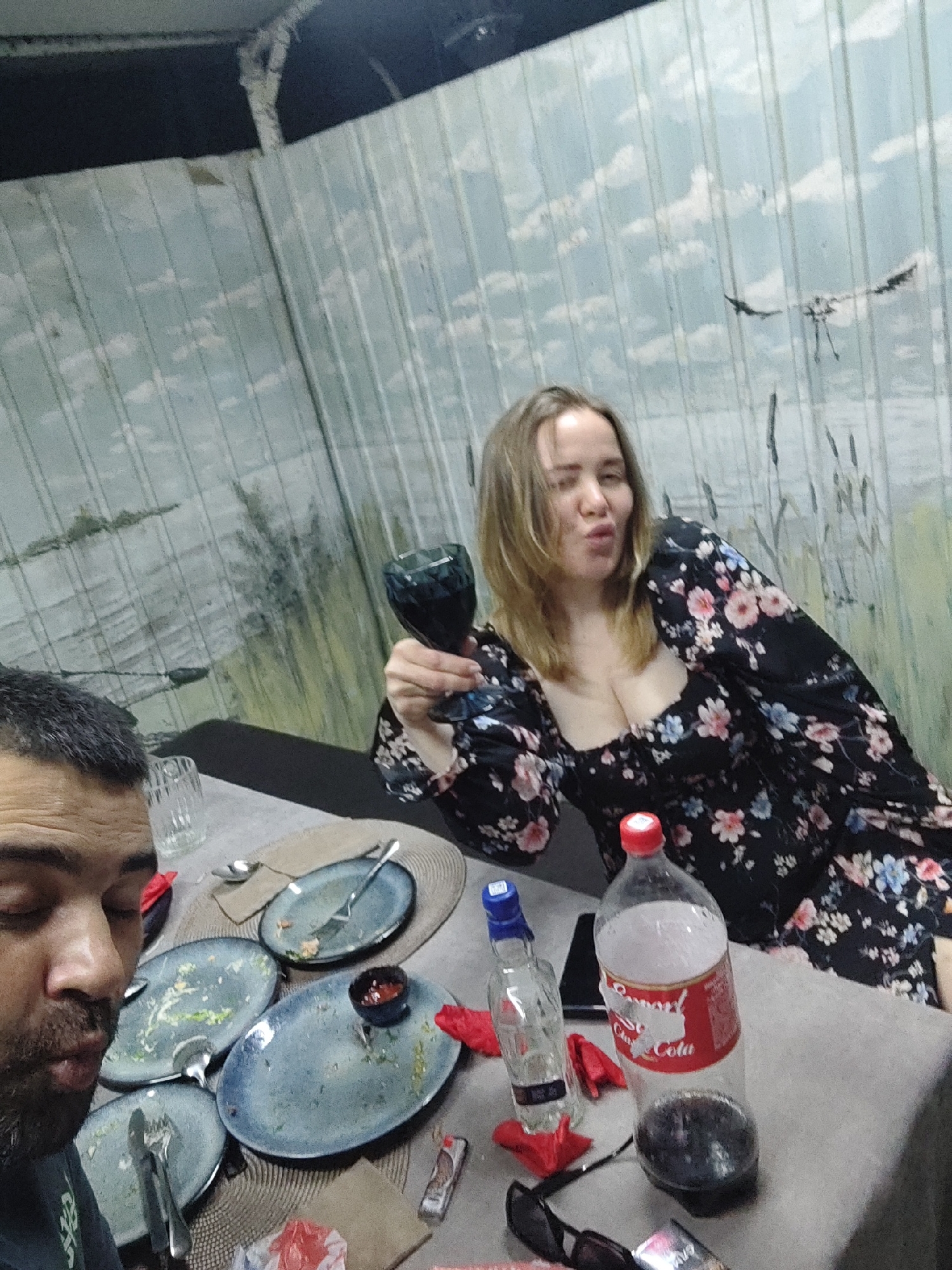 In Shimanovsk, Russia, two individuals are seated at a table, likely in an outdoor or enclosed patio setting given the painted wall depicting a cloudy sky with birds. In the foreground, a man with dark hair and beard is partially visible, his face close to the camera. He appears to be mid-conversation or in the process of eating, with his mouth slightly open. The table is covered with a woven placemat, and several dirty plates with remnants of food are scattered around. Utensils, including forks and spoons, are resting on or beside the plates. A small bowl with red sauce is also present.
To the right, a woman with blonde hair is seated, holding a dark glass in her right hand, which she is raising as if to toast or drink. She is wearing a black floral-print dress with puffy sleeves. Her facial expression is playful, with her lips pursed.
On the table, near the woman, are a nearly empty bottle of Coca-Cola with a red cap and a partially filled bottle of clear alcohol with a blue cap. Red napkins are also visible, along with a pair of sunglasses and a small lighter. The overall scene suggests a casual meal or gathering that has recently concluded or is in progress. The background wall, with its painted mural, adds a unique, if slightly faded, aesthetic to the environment.