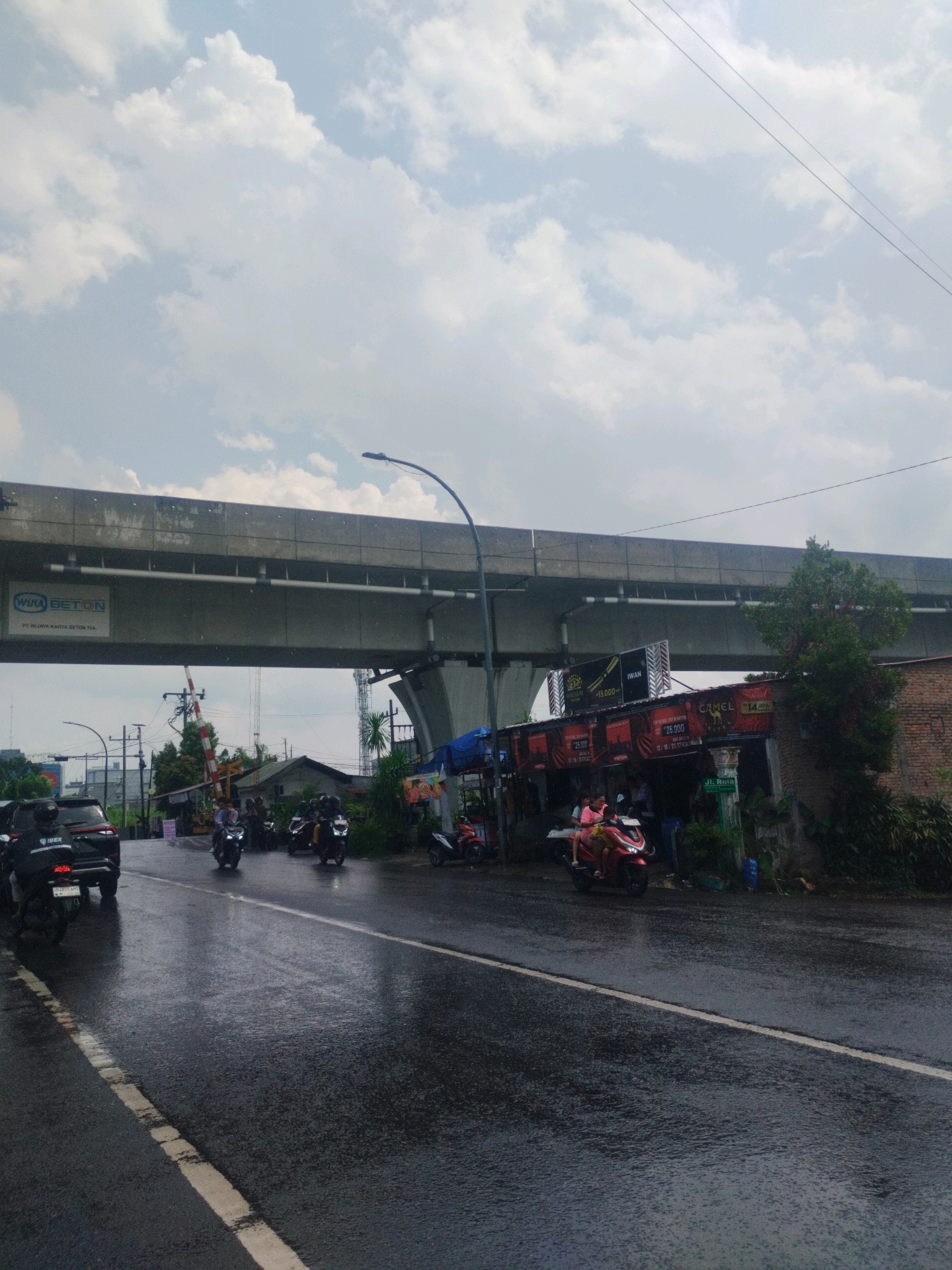 The image captures a street scene in the city of Medan, Indonesia, on a cloudy and wet day. An elevated concrete structure, likely part of a highway or bridge, dominates the upper portion of the frame. Below this structure, a road surface glistens with rain, indicating recent precipitation. Several motorcycles and a black SUV are present on the road. On the right side of the road, a row of small buildings or stalls are visible, one with a sign displaying "CAMEL" and a price. A red motorcycle with two people is stopped in front of one of these stalls. Further down the road, a railroad crossing gate is partially visible. The sky is filled with scattered clouds.