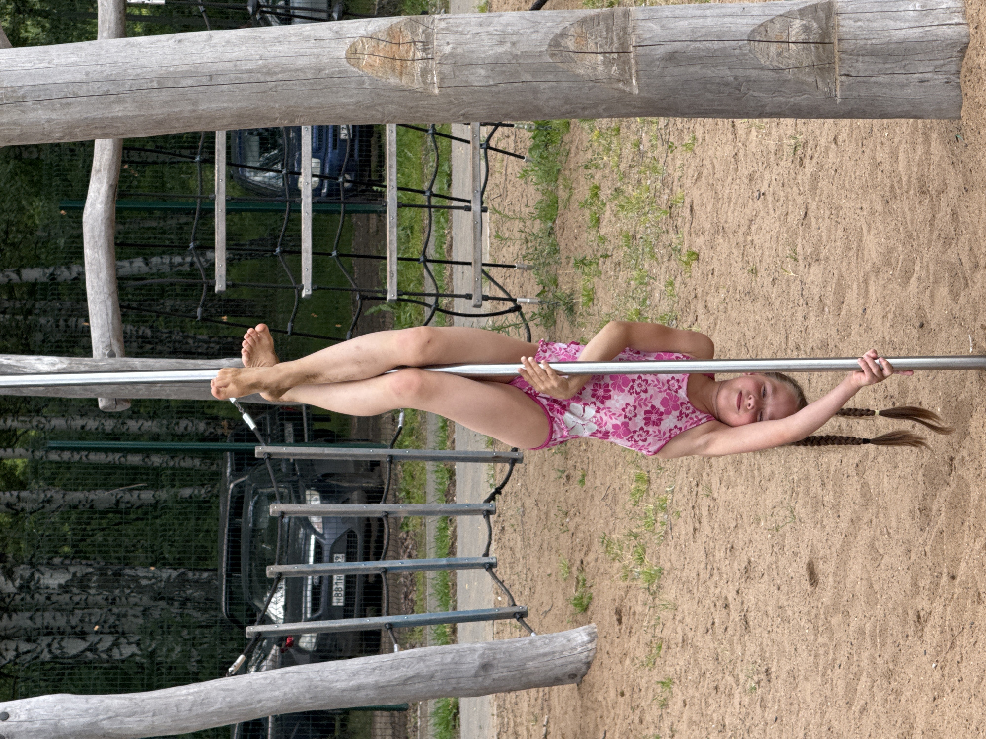 A young girl with blonde hair in braids, wearing a pink floral swimsuit, lies horizontally on a silver bar. Her body is suspended above a sandy surface, with her feet bare and her arms outstretched, one holding the bar above her head and the other extended towards her left. She appears relaxed, with her eyes closed and a gentle smile on her face. The background features a playground structure made of weathered wooden logs and metal bars, with ropes and netting visible. Behind the playground, a dense forest of birch trees can be seen, along with a dark-colored car. The sandy ground is scattered with a few blades of grass. The lighting suggests it is daytime, likely a warm, sunny day, given the clear visibility and lack of shadows indicating overcast conditions. The scene takes place in a park or outdoor recreation area, possibly a sports ground or a children's playground in Chaikovsky, Russia. The car has a license plate with Cyrillic characters, reading "H881HA."