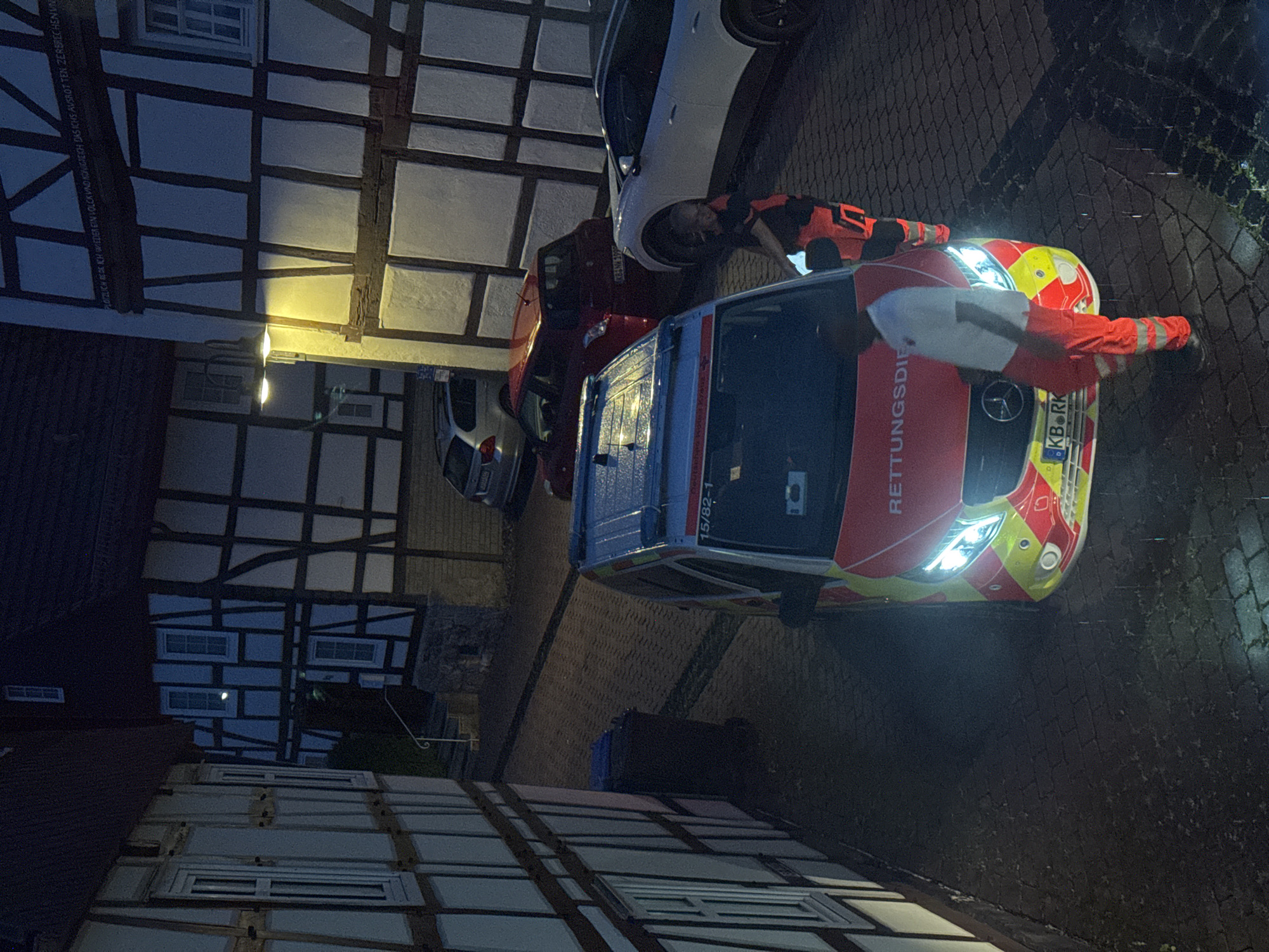 In an urban alleyway at night, an ambulance with its headlights on is parked on a cobblestone street. The ambulance is red and yellow with the text "RETTUNGSDIENST" visible on its side. Two individuals in orange and white uniforms, likely paramedics, are standing near the ambulance. One is partially obscured by the vehicle, while the other is facing the camera. The alley is lined with timber-framed buildings, characteristic of traditional European architecture. A blue trash bin is visible to the left of the ambulance. The scene appears to be in the city of Unknown, Unknown.