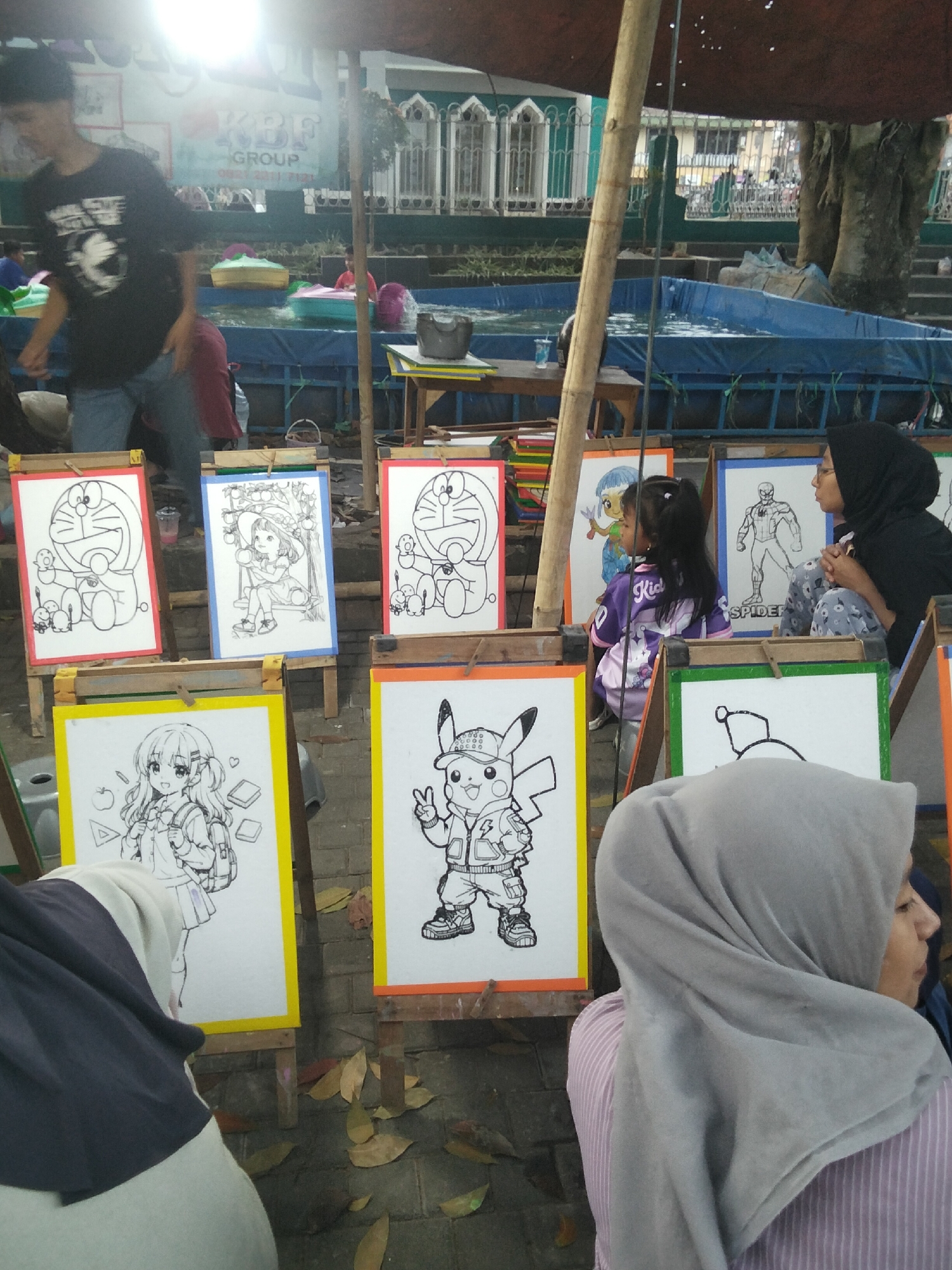The image captures a scene at an outdoor art stall in an unknown city. The focal point is a series of easels displaying black and white drawings of popular cartoon characters, including Doraemon and Pikachu, as well as anime-style characters and Spider-Man. Several people are present, including a young boy in a black t-shirt and jeans, a young girl in a purple shirt, and two women wearing hijabs. The art stall appears to be part of a larger event, with a blue tarp pool in the background, suggesting a public gathering or market. The ground is paved with bricks and scattered with fallen leaves.
