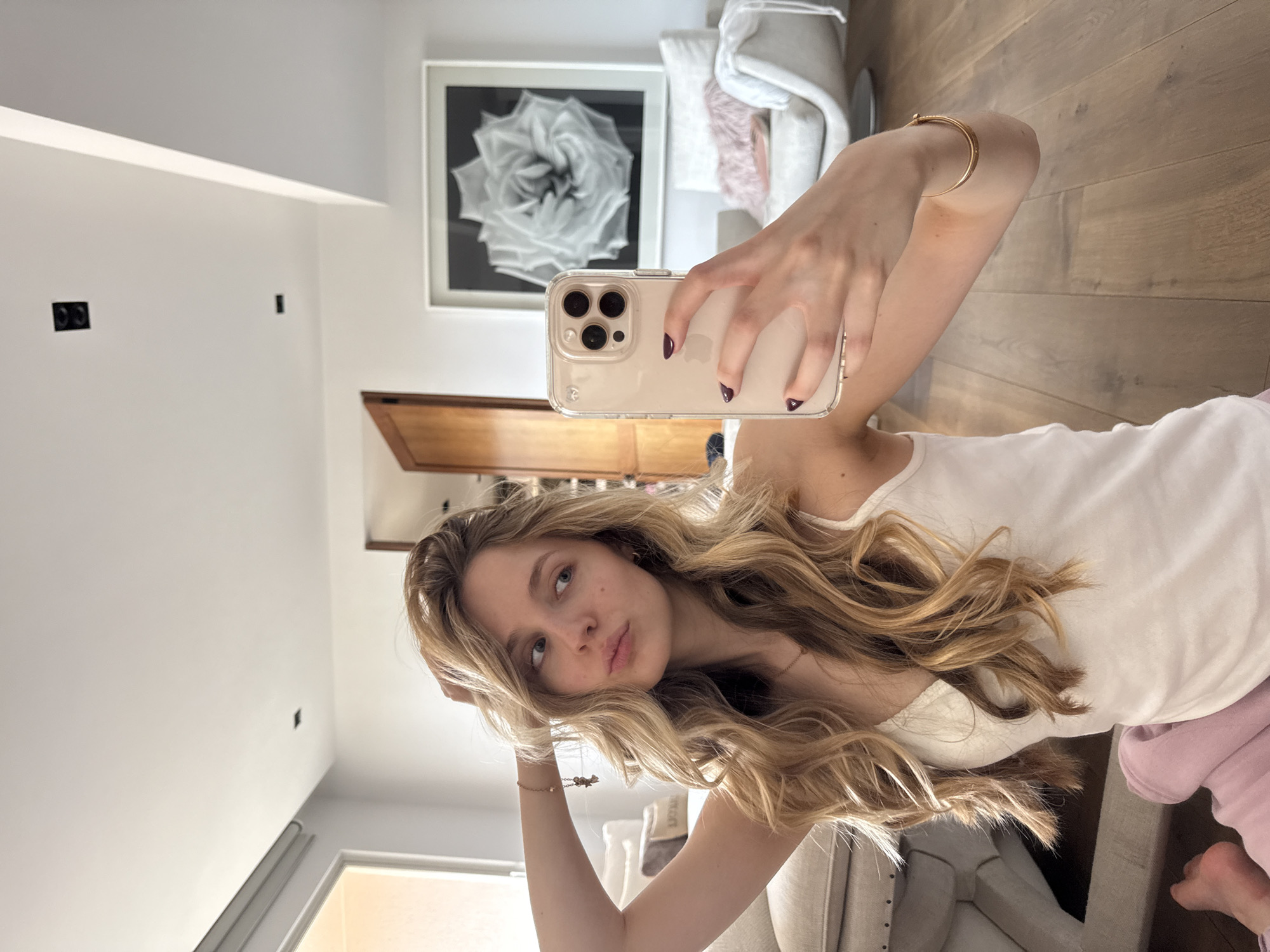 A young woman with long, wavy blonde hair is taking a mirror selfie in a well-lit indoor setting. She is wearing a white tank top and appears to have light pink bottoms. Her right hand holds a smartphone with a clear case, its triple-lens camera visible in the reflection. Her left hand is raised, touching her hair near her head, creating a casual pose. She has a relaxed expression with slightly parted lips. Her fingernails are painted a dark shade of purple or burgundy, and she wears delicate gold-colored jewelry, including a bracelet and a necklace.
The background reveals a modern room with white walls and light wooden floorboards. Prominent decor includes a large framed black and white abstract floral print, resembling a rose, displayed on the wall. Sections of light-colored furniture, specifically an armchair or sofa with a fluffy pink pillow, are also visible. Black electrical outlets and a small black rectangular object are mounted on one of the white walls. Natural light illuminates the scene, suggesting a daytime capture. The image conveys a casual, personal moment within a stylish residential interior, consistent with the provided location context of Mexico City.