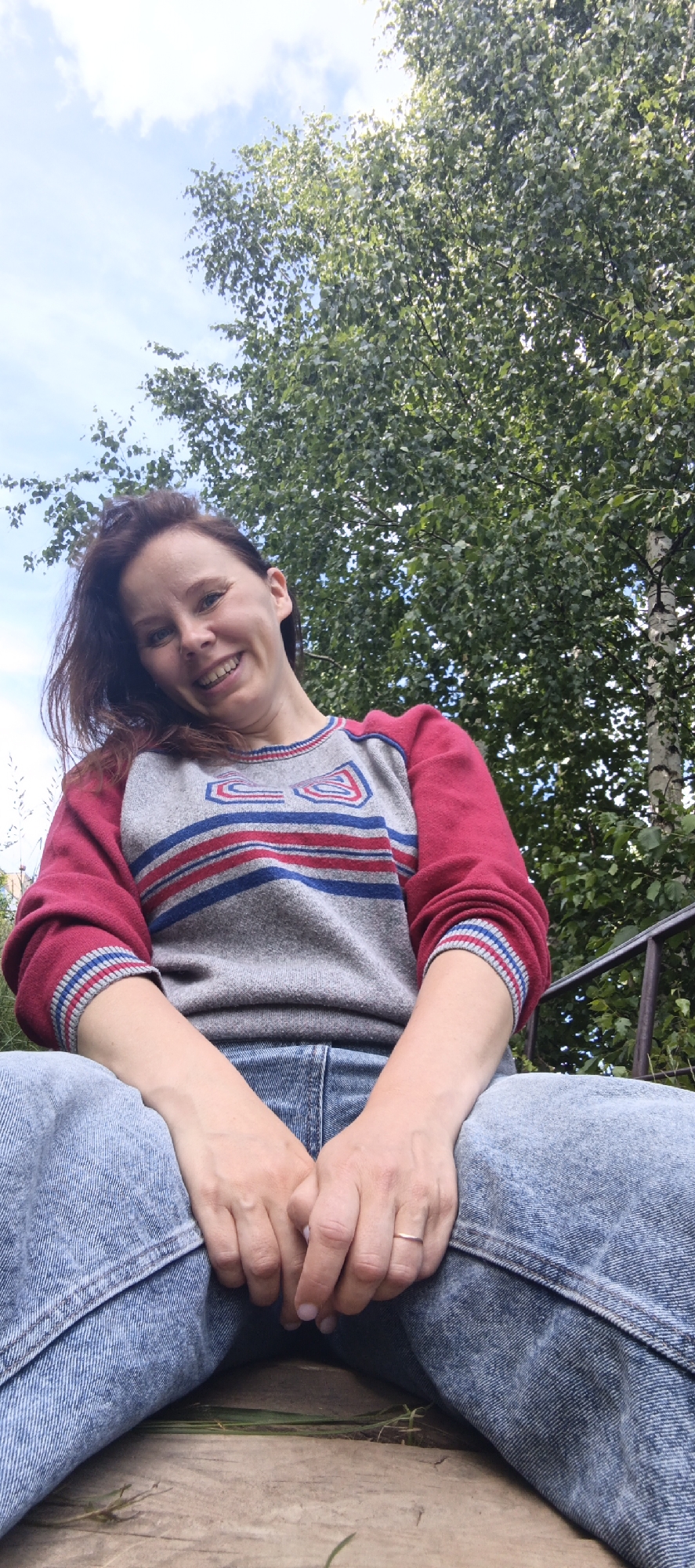 A woman is sitting outdoors, looking up at the camera and smiling. She has long, dark brown hair and is wearing a gray sweater with red sleeves and a red, white, and blue stripe pattern. She is also wearing blue jeans. Her hands are resting in her lap, and she has a ring on her left ring finger. The background consists of lush green trees and a cloudy blue sky, suggesting it is daytime and likely warm weather. The low-angle shot emphasizes the woman and the natural setting. The scene appears to be in Chaikovsky, Russia.