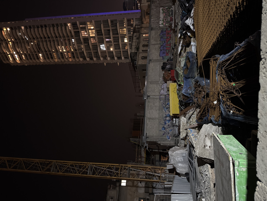 A night shot features a tall, illuminated building with numerous windows to the left. Adjacent, a dilapidated construction site is visible. This area contains graffiti-covered walls, construction materials, and debris. A crane arm stretches across the bottom of the frame, adding to the industrial, unfinished aesthetic. The scene is dimly lit, with the building on the left providing the primary light source.