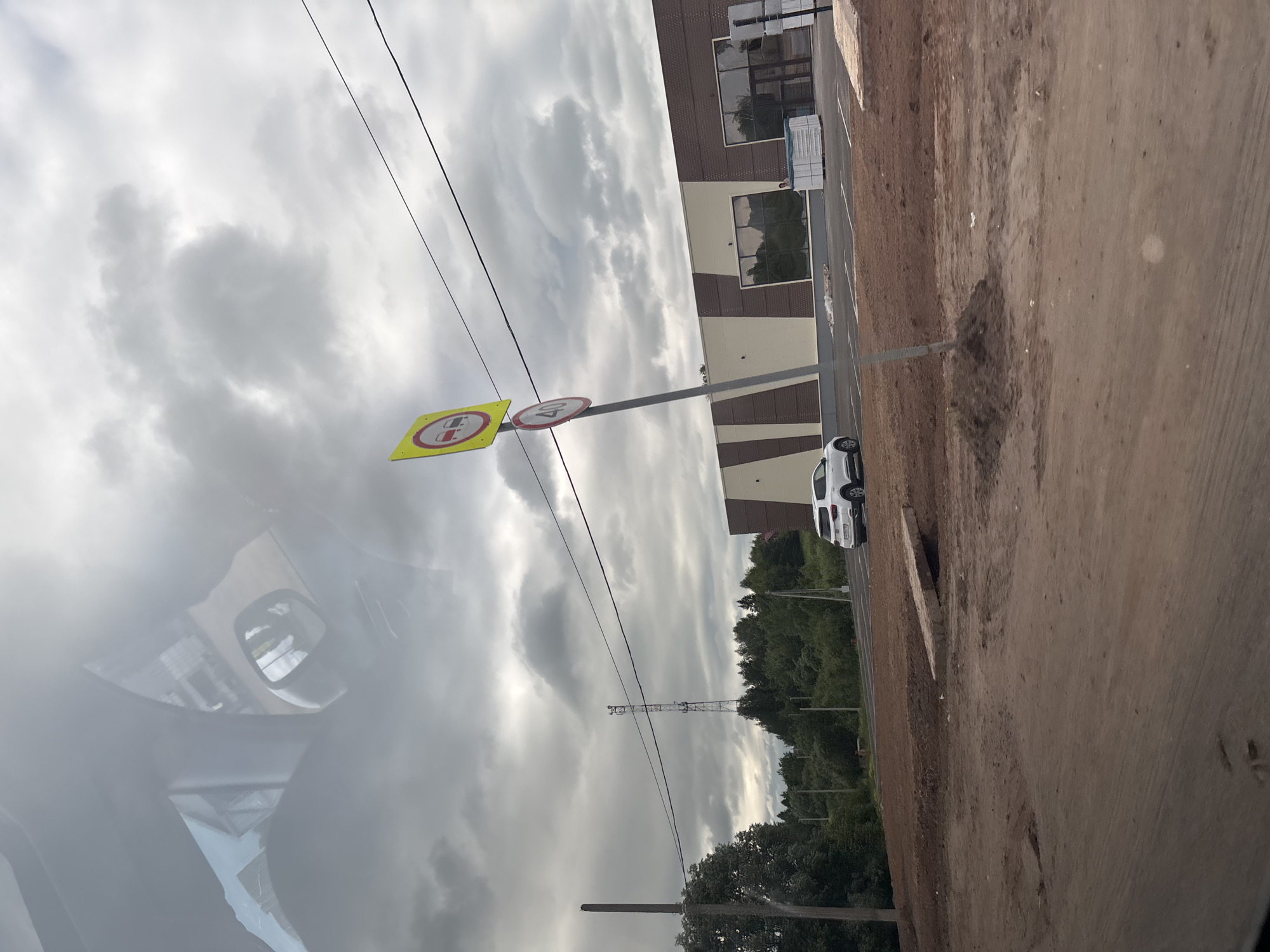 This image depicts an outdoor scene with a cloudy sky. In the foreground, there is a dirt construction site. A white SUV is parked in front of a modern building with a beige and brown facade. The building appears to be a commercial establishment, possibly a store, with large windows and a glass entrance. Two traffic signs are visible on a pole: a yellow sign with red circles indicating "no overtaking" and a circular red sign with "40" inside, signifying a speed limit of 40. In the background, a cell tower stands against the overcast sky, surrounded by trees. Power lines traverse the upper portion of the image. The scene appears to be located in the vicinity of Гаревая, Russia.