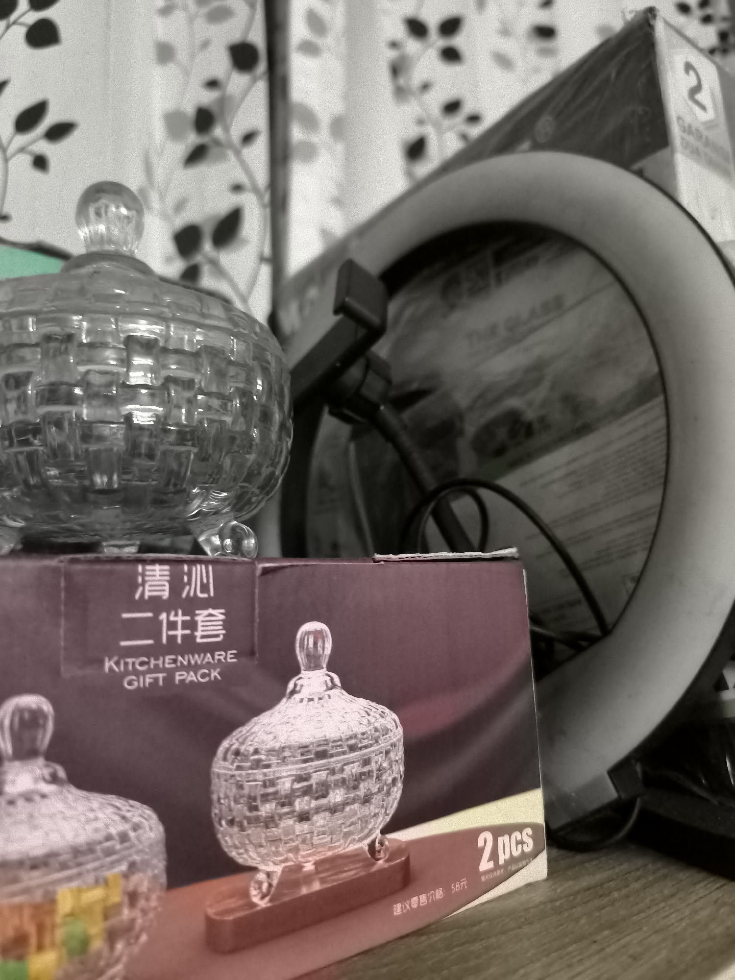 An overhead, slightly angled close-up shot shows two decorative glass bowls with lids sitting on a dark purple box. The bowls are ornate, with a woven glass pattern and small glass feet. One bowl is in the foreground on the left and contains colorful, square candies. The other bowl is slightly further back and to the right. The box in the foreground has Chinese characters that translate to "Qing Fan" and "Two-piece set" and "Kitchenware Gift Pack." To the right of the box is a large, circular ring light. Behind the ring light is a box with the number "2" prominently displayed, and the word "GARAN..." partially visible. The background consists of a curtain with a subtle leaf pattern. The lighting is dim, creating shadows.