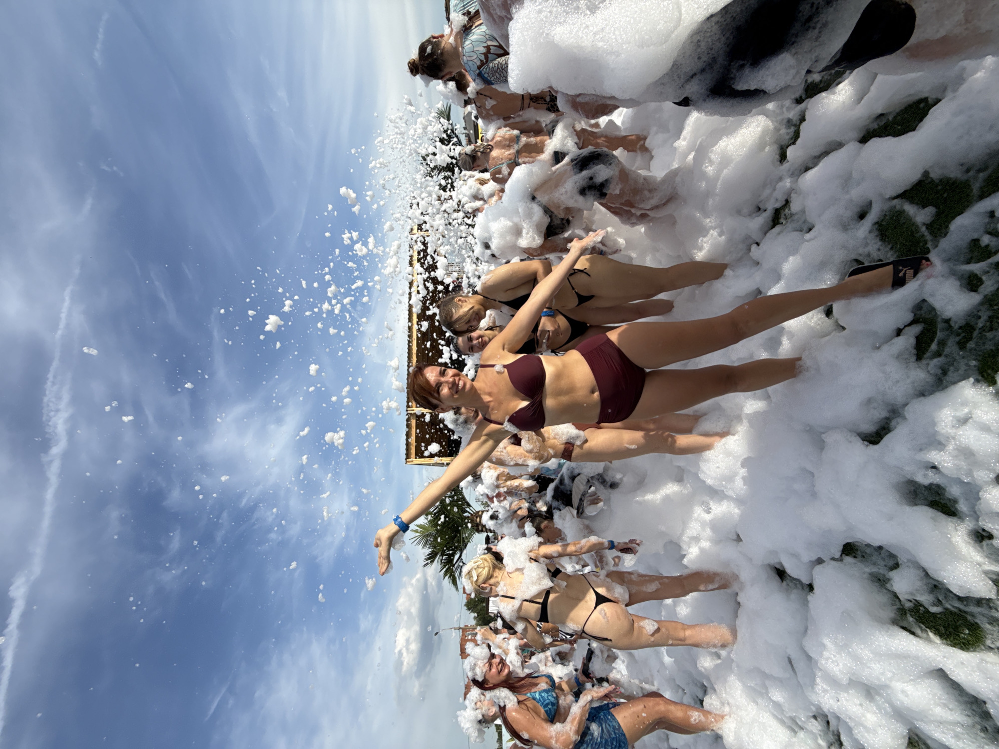 The image depicts a foam party with a group of young women in swimwear amidst a large amount of foam. The setting appears to be outdoors, possibly a beach or a venue with a grassy area. The sky is visible with clouds, and there is a structure in the background that resembles a stage or a bar. Several women are actively engaged in the foam, some smiling and posing, while others are partially obscured by the foam. The main subject in the foreground is a woman in a burgundy bikini, with her arms outstretched and a joyful expression. The overall atmosphere suggests a lively and celebratory event. There is no indication of the location being the city of Rusanovo, Russia.