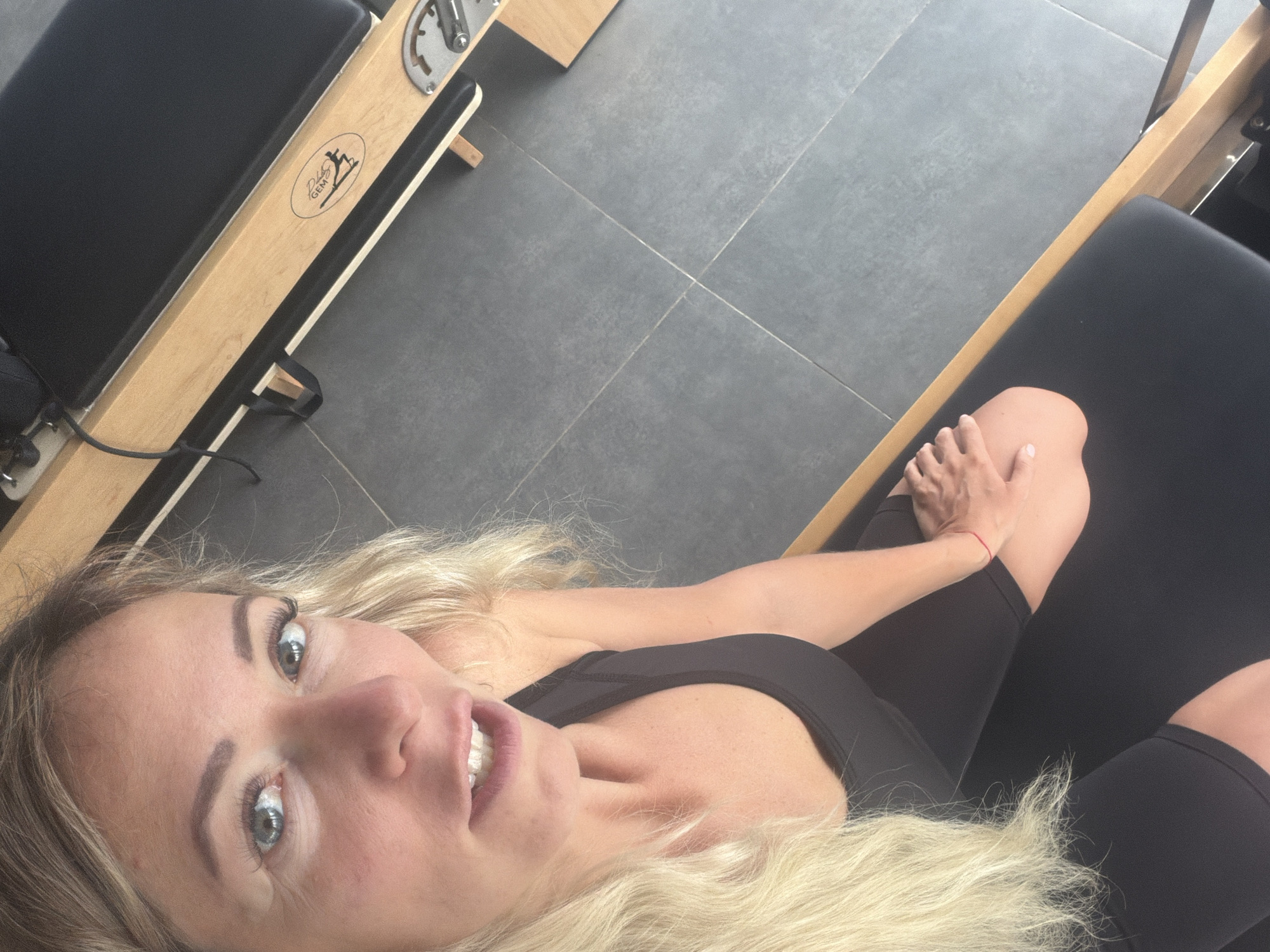 A woman with blonde hair and blue eyes is taking a selfie in a room with grey tiled flooring. She is wearing a black tank top and black leggings. She is sitting on a black mat which is part of a wooden Pilates reformer. The Pilates reformer has a logo on its side that appears to say "Body Gem". The lighting is bright, suggesting the photo was taken during the day. The woman's right hand is resting on her left knee. There is a thin red string bracelet on her right wrist.