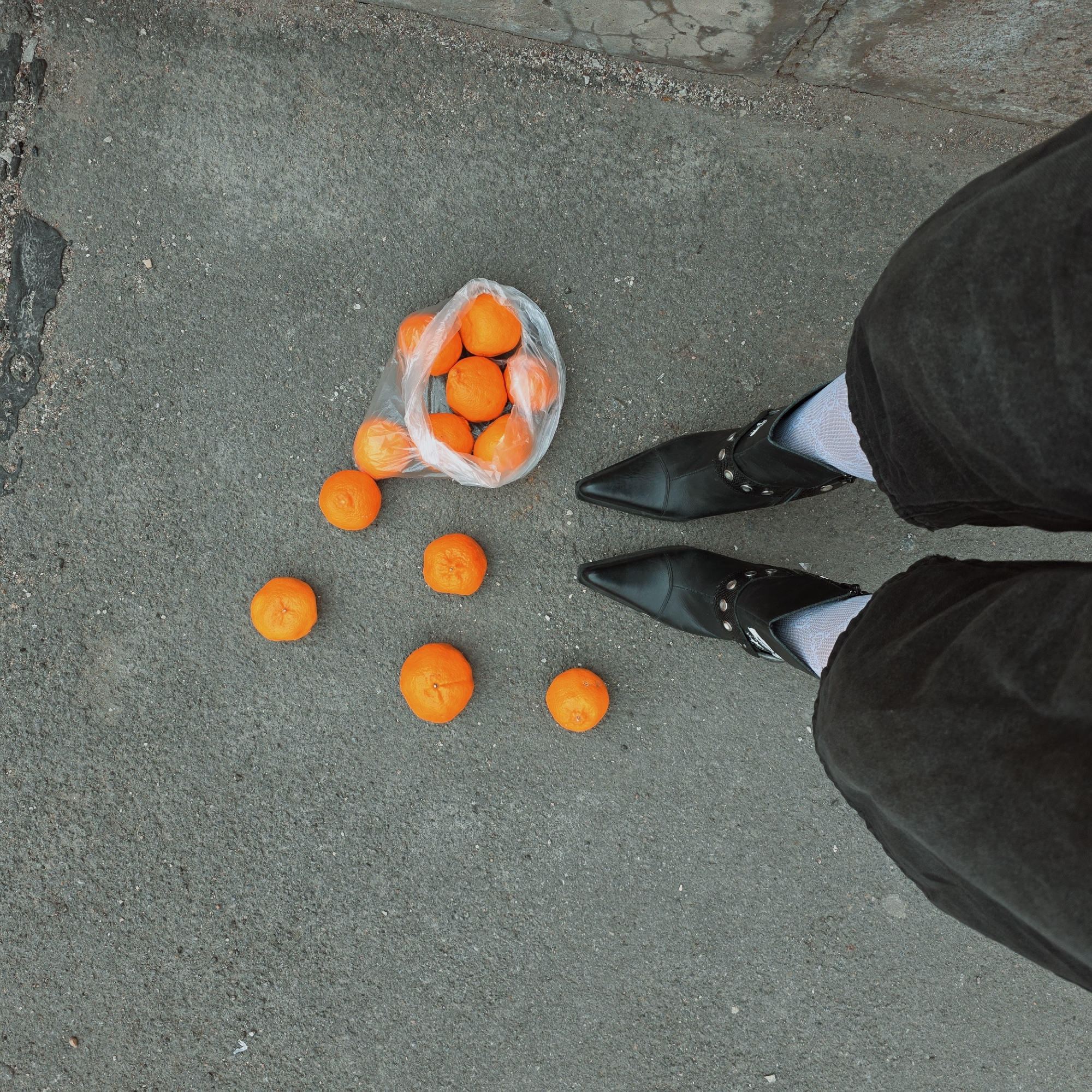 A high-angle shot shows a person standing on gray asphalt, possibly in a city, with tangerines scattered around. The person wears black, pointed-toe boots with silver accents and white patterned socks, and dark denim or canvas pants. A clear plastic bag containing more tangerines sits near the person's feet, and several tangerines have spilled out onto the asphalt. The edges of the pavement are visible in the upper corner of the picture. Based on the style and architecture that I am trained on, this location appears to be in Saint Petersburg, Russia.