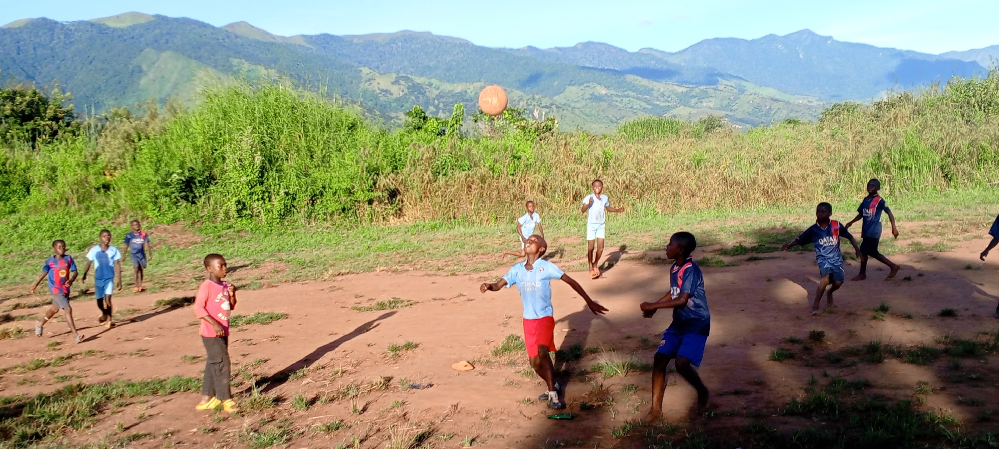 In a daytime outdoor shot taken in Ngalula, Democratic Republic of the Congo, a group of approximately ten children are playing with a ball on a dirt field. The field is surrounded by green vegetation and tall grass, with mountains visible in the background.
The children are of African descent and appear to be boys, wearing casual clothing like t-shirts and shorts. One boy is wearing a pink t-shirt and dark pants with yellow shoes. Several children wear sports jerseys. The ball is in mid-air, suggesting an active game or play session.
The environment includes a clear sky and sunlight, indicating a sunny day. There are shadows of trees and figures on the ground. The terrain is uneven and rustic.