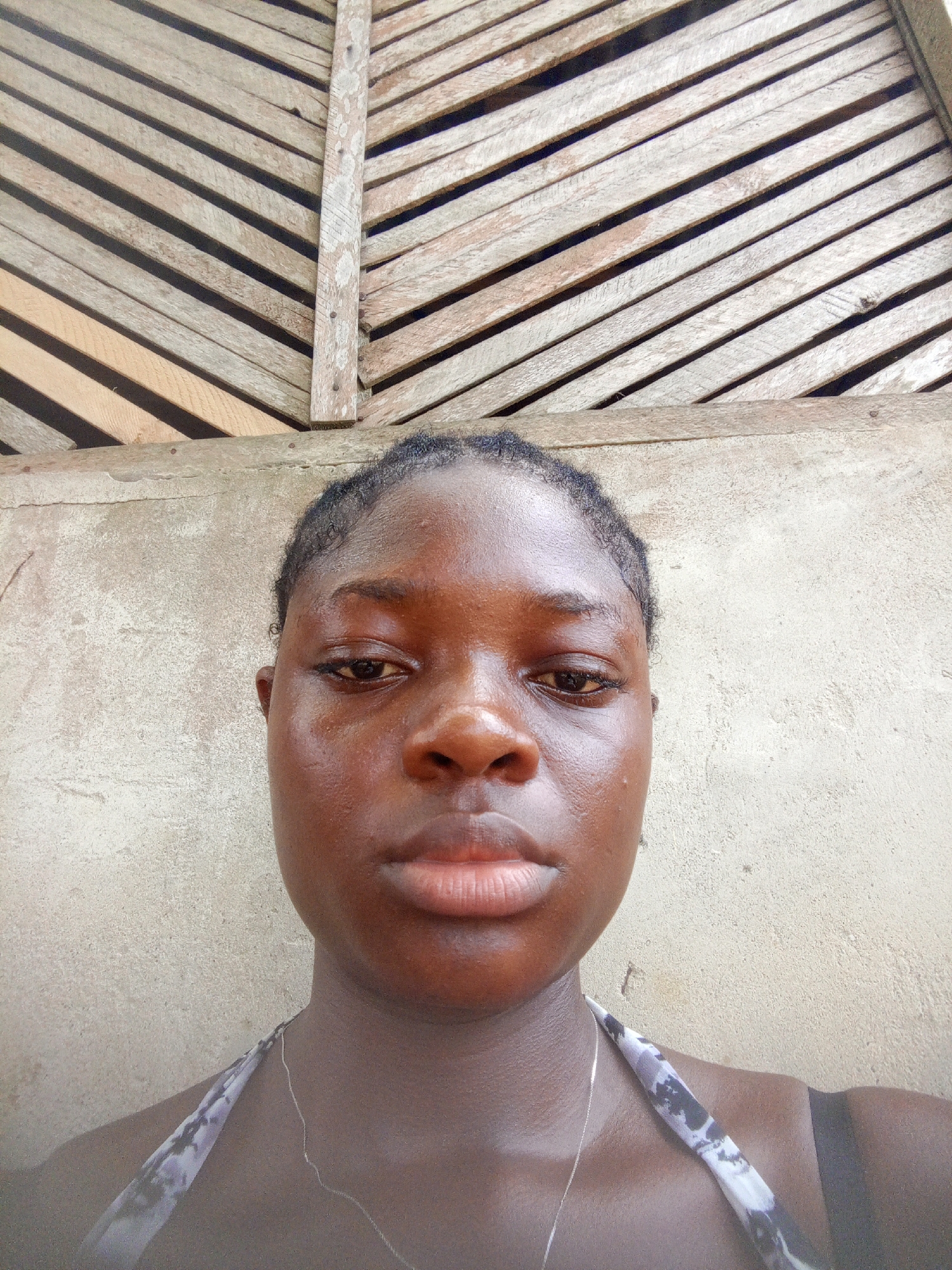A close-up of a young woman with dark skin. Her hair is braided and pulled back from her face. She is wearing a silver necklace and a tank top with a black and white print. Her expression is neutral. Behind her is a rough, light-colored wall and a wooden structure with slats. Based on the prompt, the location is in the city of Mbribit, Nigeria.