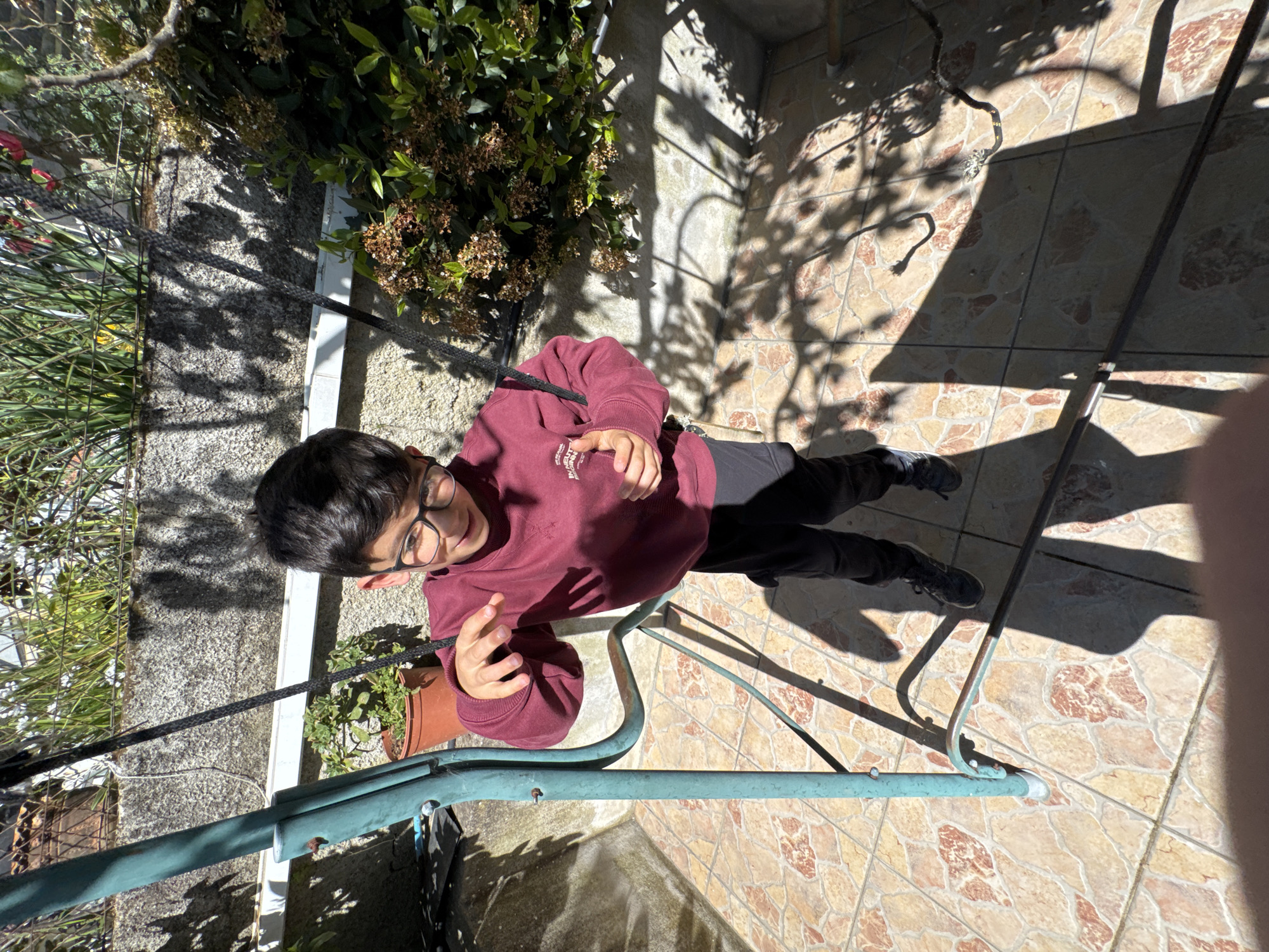 The scene depicts an outdoor residential area in the city of Mirandela, Portugal, on a brightly lit day. A young male, approximately 8-12 years old, with dark hair and wearing dark-rimmed glasses, stands center-frame. He is dressed in a maroon long-sleeved sweatshirt with faint white text on the left chest, dark pants, and dark athletic shoes. His left arm is raised with the hand open, fingers slightly splayed, while his right arm is bent with the hand near his chest. He is looking down and to his right, with a discernible expression on his face.
The ground consists of patterned ceramic tiles in shades of tan and reddish-brown. Strong sunlight casts distinct shadows across the tiled surface, including the subject's elongated shadow and shadows from overhead structures or foliage.
To the left, a teal-colored metal frame, possibly part of a swing set or garden furniture, extends vertically and then horizontally. Some rust is visible on the horizontal section of this frame. A dark, thin cord or rope hangs vertically near the left side of the frame. In the background, a concrete wall topped with a metal wire fence is partially visible. Green foliage, including a plant in a terracotta pot, is positioned behind the wall. To the right of the subject, a dense green bush with clusters of dried, light-brown flora is visible.