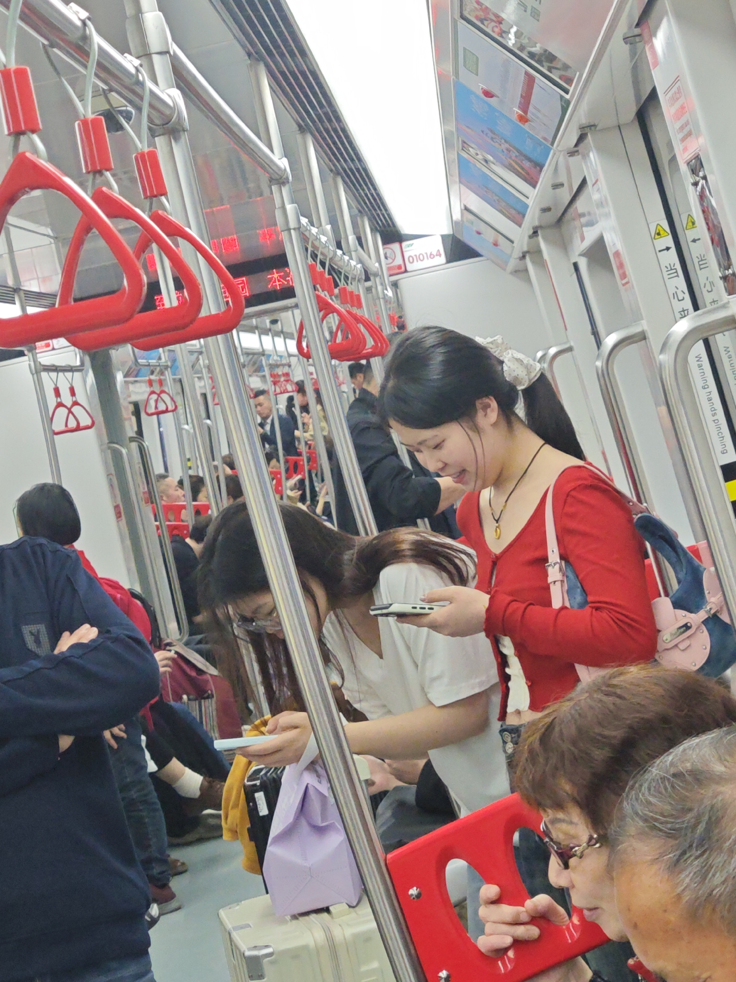 An indoor scene inside a metro rail car in Shapingba District, China, depicts several people. A young Asian woman in a red long-sleeved shirt and a blue purse looks at her smartphone, smiling. Adjacent to her is another young Asian woman in a white t-shirt, also looking down at her smartphone. In the foreground, an elderly woman with glasses holds onto a red grab handle. The background shows numerous other passengers, luggage, and the interior of the train car with red grab handles and stainless steel poles.