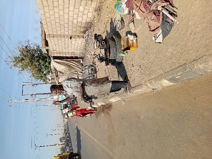 The image depicts a daytime outdoor scene in Sharada, Nigeria, alongside a paved multi-lane road under a clear blue sky. An adult male, of apparent African descent, stands on an unpaved shoulder bordering the road in the foreground. He wears a heavily soiled light-colored tunic over dark trousers and sandals. His right hand is raised with an open palm, and his left hand holds a dark, elongated object.
Adjacent to him on the ground, mechanical components are visible: a small engine mounted on a frame rests on two dark-colored tires. Nearby are a yellow plastic crate, a green plastic bottle, a metal bowl, and assorted piles of fabric and debris. To the right of the scene, a partially constructed light-colored block wall and a makeshift structure covered with a light-colored tarp are situated. A tree with green foliage is visible behind these structures. Overhead utility lines and poles run parallel to the road.
In the mid-ground on the paved road, a second individual, seen from behind, walks away from the viewer. This person is dressed in red clothing, overlaid with a yellow reflective vest or harness, and appears to carry a light-colored bag or object in their left hand. On the far left of the road, a yellow dump truck is partially visible, moving away from the camera. Additional vehicles and modern streetlights are discernible in the distant background, indicating an urban or semi-urban roadway.
