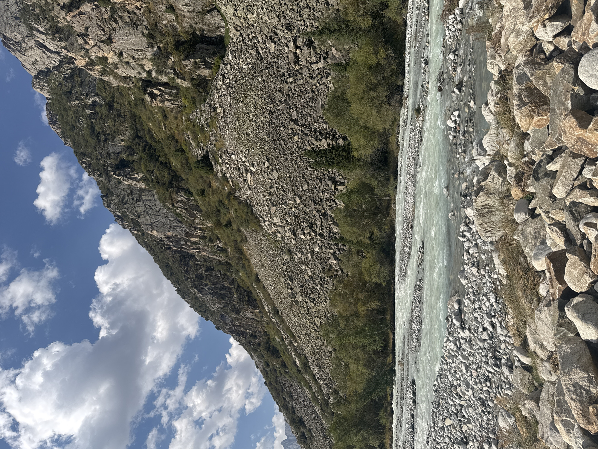 An eye-level, long shot captures a mountainous landscape under a bright, partly cloudy sky in an unknown location in Kyrgyzstan. The dominant feature is a steep, rocky mountain slope covered in scree and sparse vegetation, with jagged rock formations and patches of green foliage. At the base of the mountain, a wide, shallow river flows from left to right across the frame. The water is milky turquoise and turbulent, with numerous gravel bars and submerged rocks visible. The foreground is occupied by a bank of large, weathered rocks and dry grasses. The scene is serene and natural, with no signs of human presence.