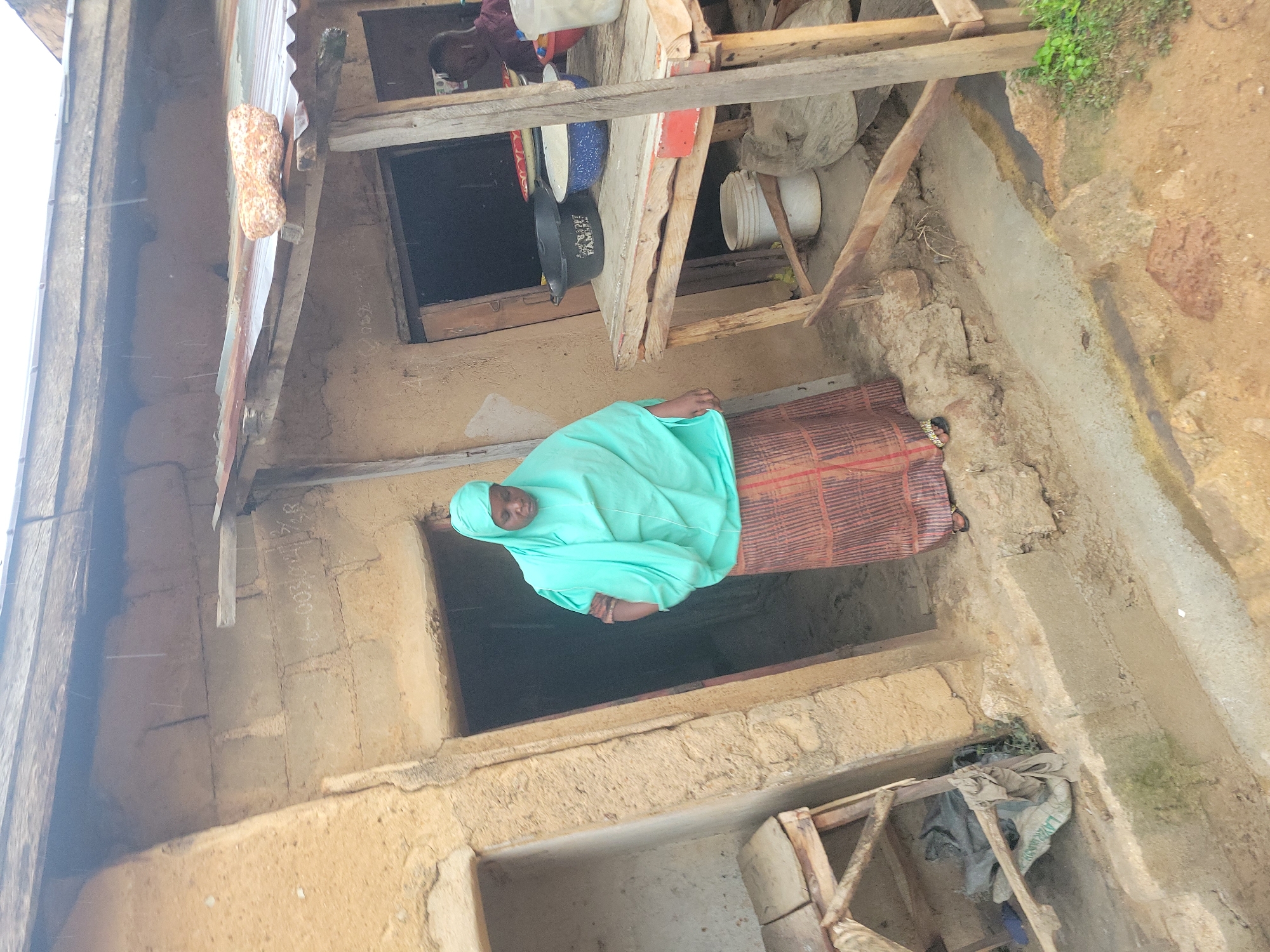 In Jos, Nigeria, a woman wearing a light blue hijab and a patterned skirt stands in a doorway of a rough, tan-colored building. The building has a corrugated metal roof and a wooden stall to the right of the doorway. On the stall are several bowls, one black with white lettering that reads "247 FAMILY," and a white bucket. Behind the woman, a dark opening suggests another room or passage. To the left of the doorway, there is an empty shelf built into the wall. The ground in front of the building is dirt and concrete. To the far right, a person is partially visible behind the stall.