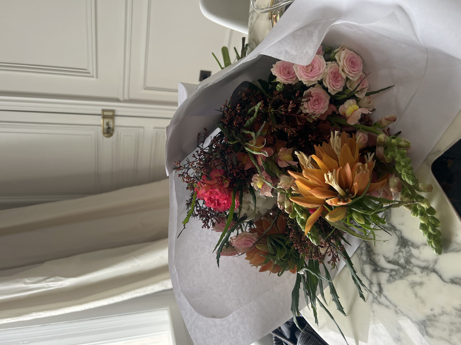 A bouquet of flowers wrapped in white paper sits on a marble surface. The bouquet contains roses, an orange flower resembling a protea, and greenery. To the left of the bouquet, a window with a curtain is visible. Behind the bouquet, there is a cabinet or wall with paneling and a small metal latch.