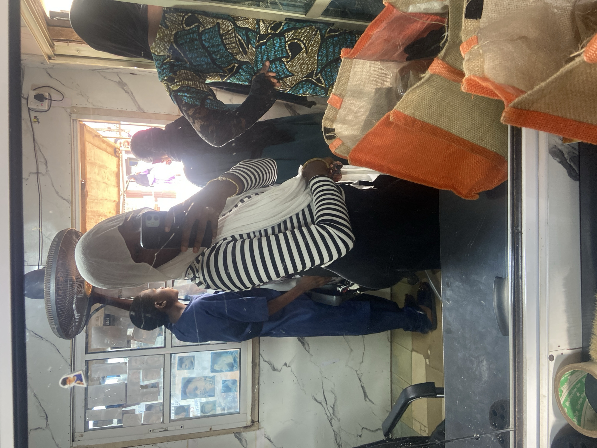 A reflection in a mirror shows the interior of a hair salon in Okengwe-Obehira, Nigeria. A woman wearing a black and white striped top and a white head covering is taking a photo with her cell phone. A person in a blue uniform is working on the hair of a seated customer. Another person with a black head covering and a patterned turquoise and black dress is standing nearby. A fan is visible on the left. The background includes a window covered with photos. Orange-trimmed burlap bags hang at the top right.