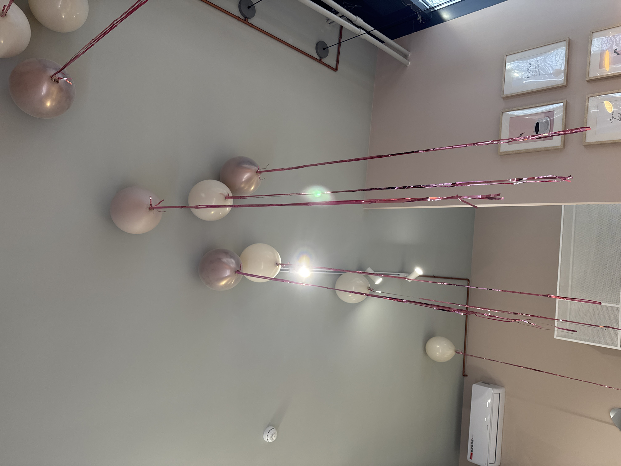 The image shows a room decorated with white and rose-colored balloons attached to pink metallic ribbons. The balloons are dispersed across the ceiling, suggesting a celebratory event. Visible in the background are decorative framed pictures on a light pink wall, pipes running along the ceiling, and an air conditioning unit attached to the wall. There are no people visible in the frame. The location is Unknown, Unknown.