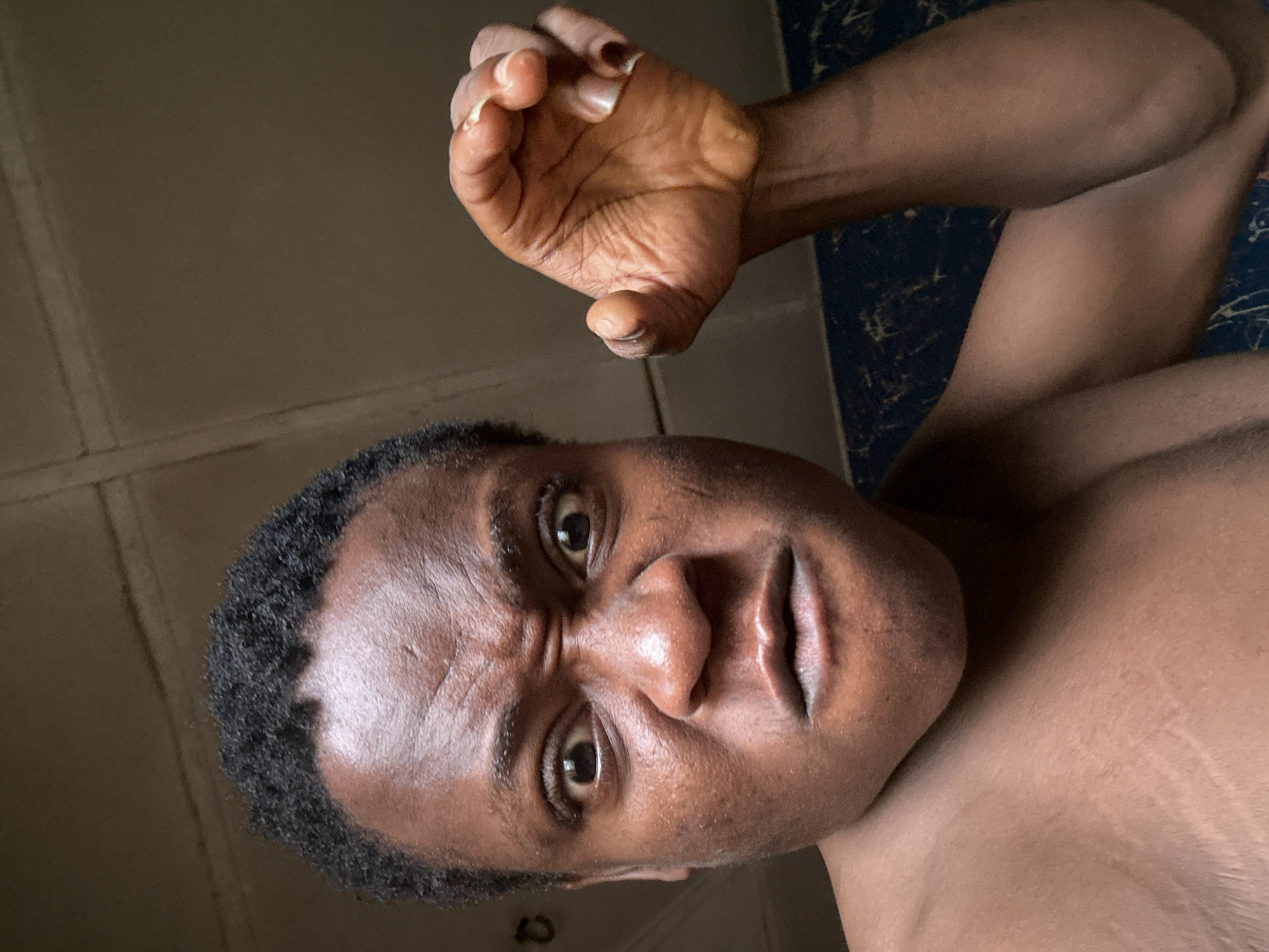 A close-up, low-angle portrait shows a shirtless Black man with short, dark curly hair. His face displays a strained expression, with furrowed brows and a slightly open mouth. His right hand is raised and clenched into a loose fist. The background consists of a tan-colored wall with horizontal lines. The location is Unknown, Unknown.