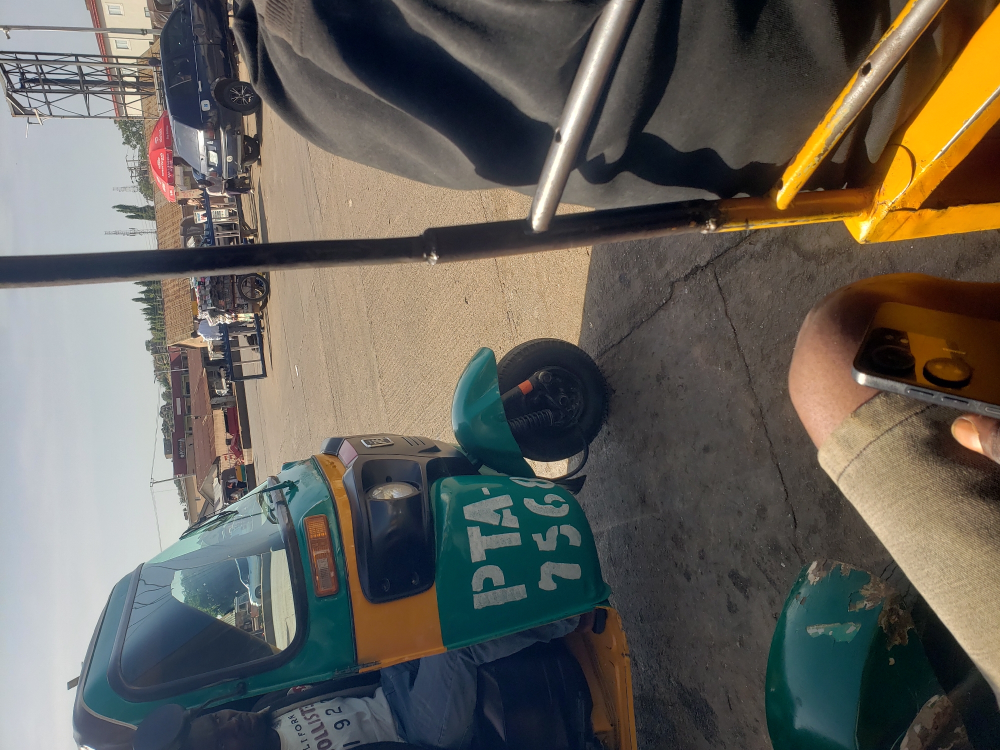 The image captures a sunny day scene at a busy street corner in Jos, Nigeria. A green and yellow auto-rickshaw, known locally as an "okada" or "keke," is prominently featured in the foreground, with white lettering on its side reading "PTA-7568". A man is seated in the driver's seat of the auto-rickshaw, looking towards the left. Another individual, partially visible, is seated behind the driver. The auto-rickshaw appears to be stationary, possibly waiting for passengers or at a traffic stop.
In the background, several other vehicles are present, including a dark blue SUV. A red umbrella is visible, suggesting a vendor or a roadside stall. Buildings line the street, with some appearing to be shops or businesses. The sky is clear and blue, indicating fair weather. The overall impression is one of a typical urban environment in Jos, with the hustle and bustle of daily life.