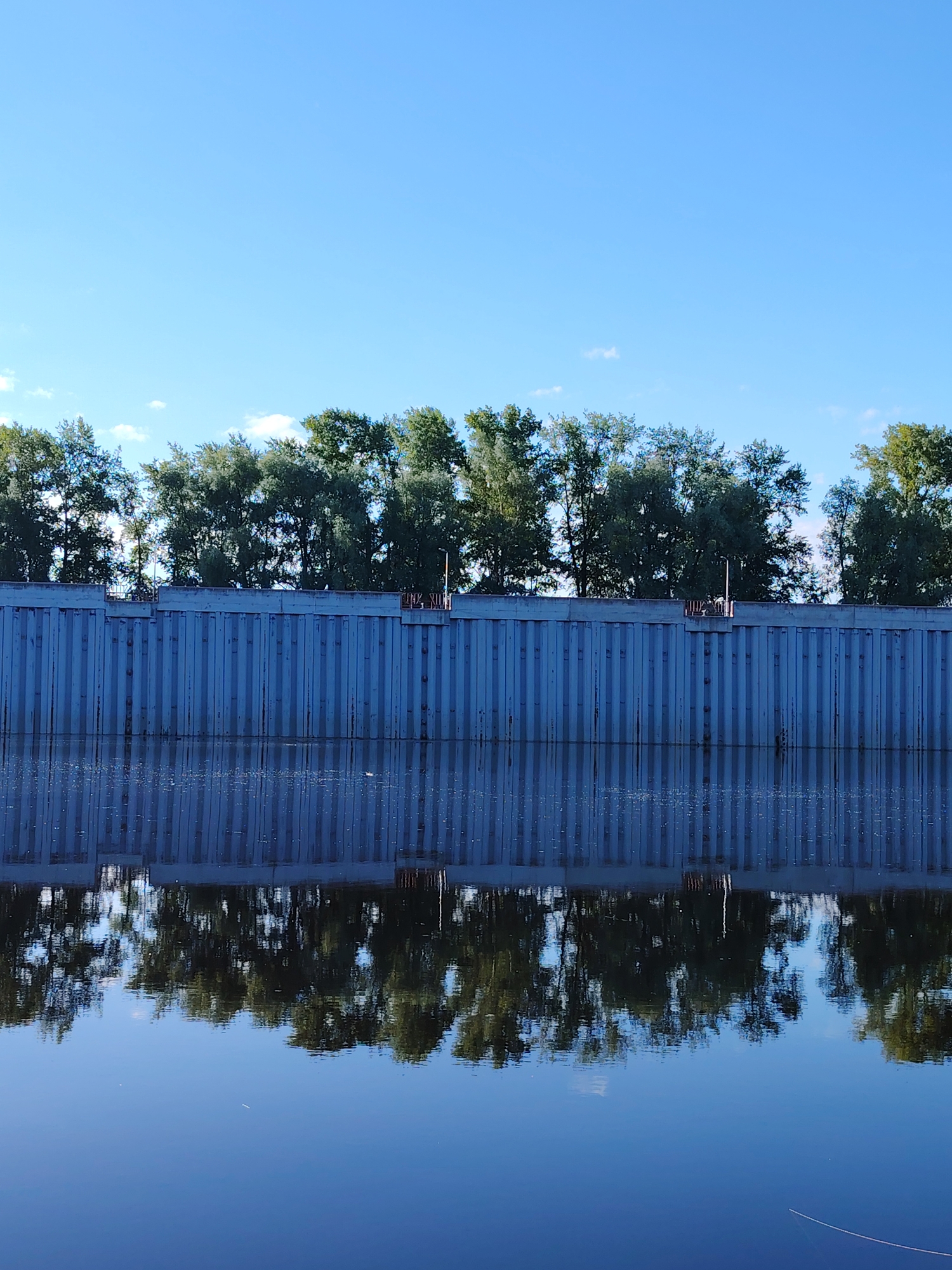 The image depicts a calm river with a reflection of a tall, blue, corrugated metal wall and a line of trees against a clear blue sky. The wall appears to be a retaining structure or barrier. The reflection is sharp and detailed, showing the textured surface of the wall and the foliage of the trees. The environment suggests an outdoor, possibly urban or industrial setting, given the man-made wall. The location is identified as the embankment of the Kama River in the city of Chaikovsky, Russia.