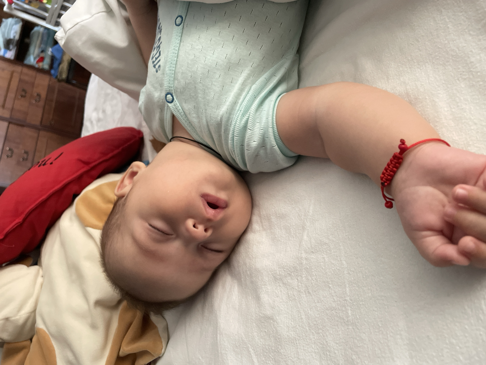 A baby is sleeping peacefully on a white bed, with their eyes closed and mouth slightly open. The baby is wearing a light blue onesie and a red bracelet on their left wrist. In the background, there is a red pillow and some drawers. The image appears to be taken indoors, possibly in a bedroom. There is no specific indication of location within Fălești, Moldova.
