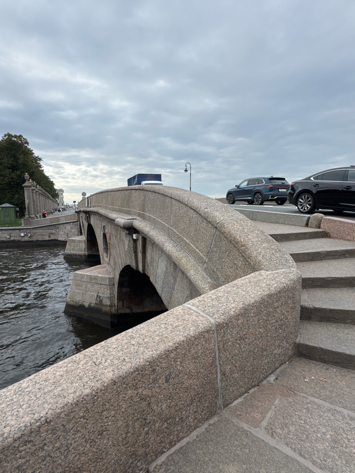 This is a medium-angle, eye-level, full shot of a stone bridge arching over a dark body of water, likely a canal or river, in Saint Petersburg, Russia. The bridge's architecture features rough-hewn granite blocks forming its structure, with a rounded parapet leading up to the roadway. To the right, a set of stone stairs ascends the bridge's embankment. The roadway on top of the bridge has two vehicles: a dark-colored sedan on the right and a dark green SUV to its left. The background reveals a cloudy, overcast sky, and on the far left, a tree-lined embankment with a row of ornate stone pillars and a small green structure. A few pedestrians are visible on the embankment path. The water appears choppy, suggesting a breeze. The overall scene depicts an urban waterway with a historic stone bridge, characteristic of Saint Petersburg's cityscape.