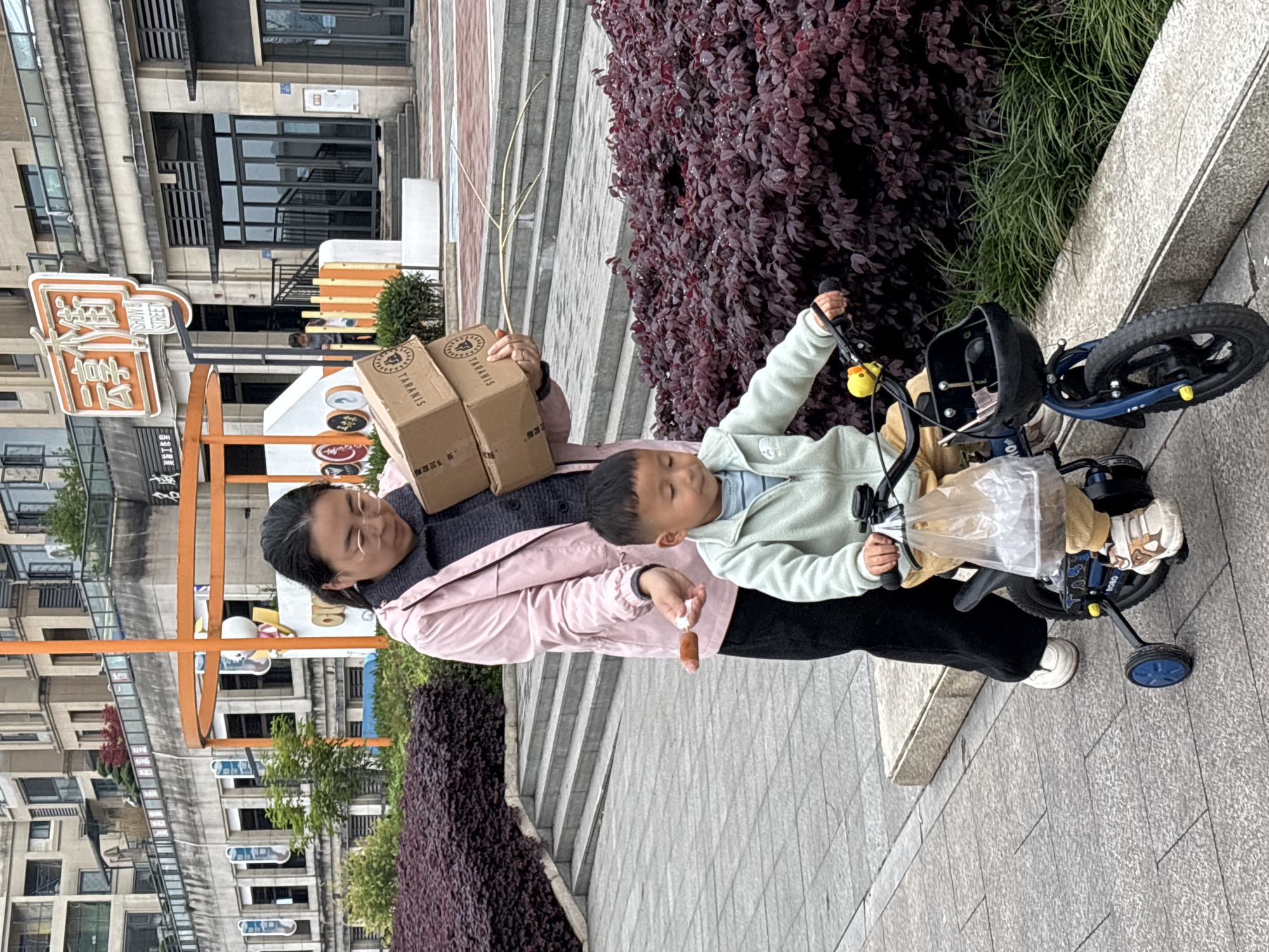 The image captures an adult female and a young child on a paved pedestrian walkway in Jiangjin, China, adjacent to a commercial street identified by a prominent orange sign displaying "云享街" (Yun Xiang Street) and "SHOW STREET." The adult female, appearing to be in her late 30s to 40s, wears glasses, a pink jacket over a dark grey sweater, and dark pants. She holds two stacked brown cardboard boxes labeled "TARANIS" in her left arm. Her right hand is extended, presenting a brown, sausage-like food item towards the child. The child, a male approximately 2-4 years old, is dressed in a light green zip-up fleece jacket and light brown pants, and is seated on a dark blue tricycle equipped with training wheels and a small front basket. A clear plastic bag is suspended from the tricycle's handlebars. Both individuals are looking downwards. The immediate foreground features dense reddish-purple leafy bushes, behind which concrete steps ascend. The background consists of multi-story buildings with windows and balconies, characteristic of an urban commercial or residential area. The ground surface is paved with grey interlocking tiles.