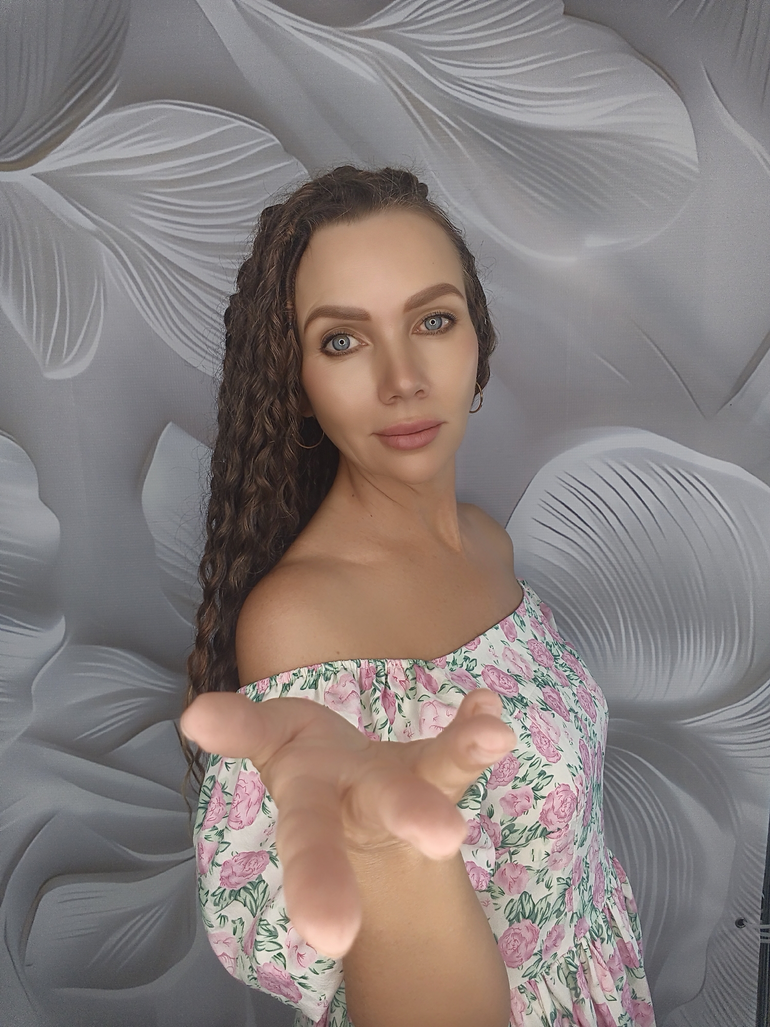 A woman with long, curly brown hair and blue eyes, wearing an off-the-shoulder floral dress, extends her right hand towards the camera. The background is a light-colored wall with a textured floral pattern. The woman is looking directly at the camera with a neutral expression. The lighting is soft, illuminating the woman's face and the foreground.