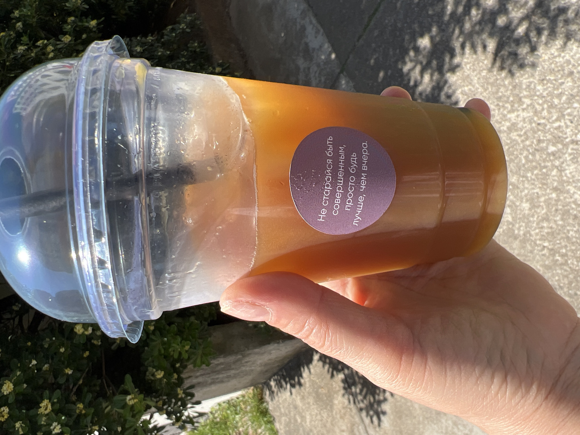 A hand holds a clear plastic cup with a rounded lid. Inside the cup is an orange liquid. A round purple sticker on the cup has white text in Cyrillic script. The Cyrillic text translates to "Don't strive to be perfect, just be better than yesterday." The background is slightly blurred, showing greenery and a concrete surface. The photo appears to be taken outdoors in daylight. Based on the sticker's language (Russian) and the context of the image, it can be inferred that the location is likely Budva, Montenegro due to the high number of Russian speakers and tourists there.