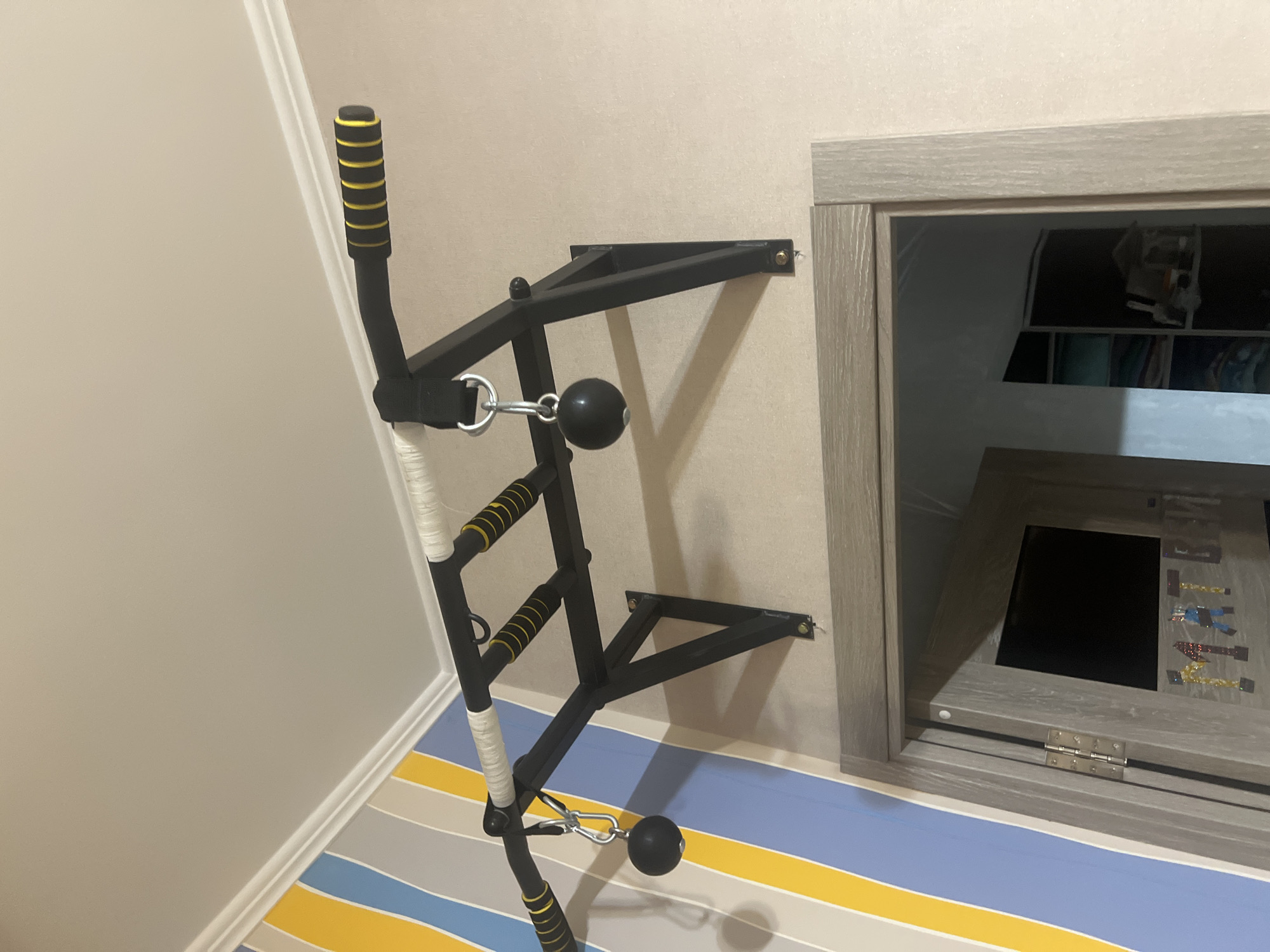 The image shows a wall-mounted pull-up bar attached to a wall with beige wallpaper. The pull-up bar is black metal with yellow-striped rubber grips and white wrapping on a central section. Two black, spherical weights hang from the bar via metal carabiners and chains. To the left of the pull-up bar, a wall is decorated with vertical stripes of blue, yellow, and gray. Below the pull-up bar, a wooden door frame is visible, with a door partially open, revealing the interior of a room with shelves and clothing. There is no discernible information regarding the city of Veteran-2, Russia, in the image.