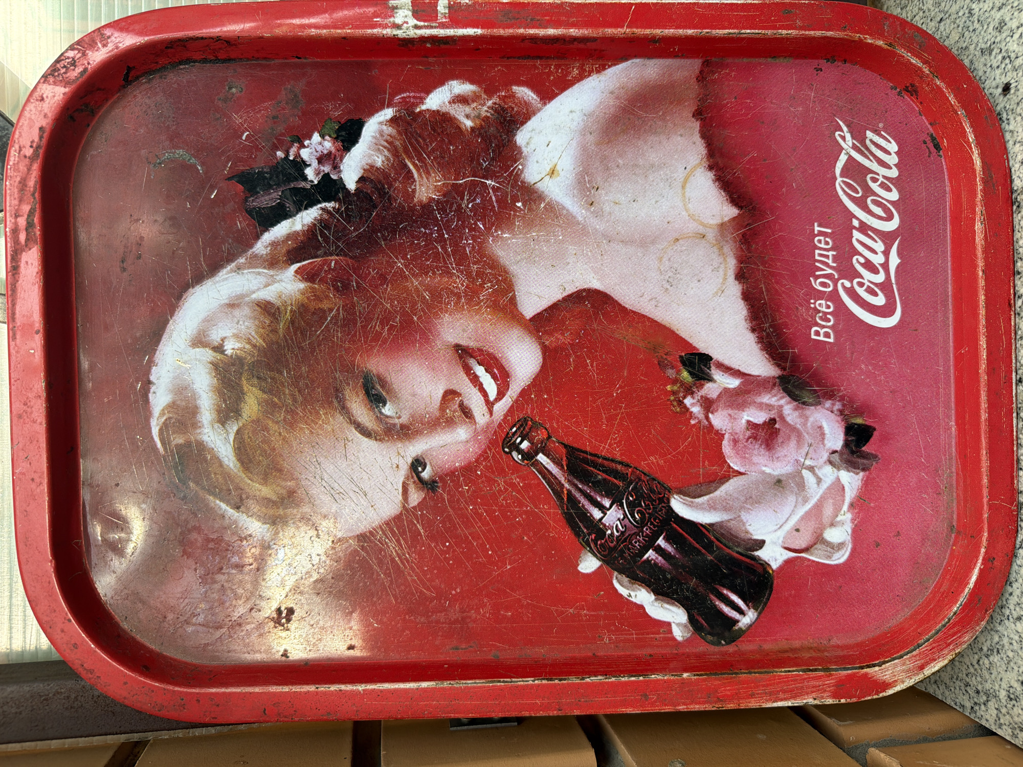 A vintage Coca-Cola tin tray is displayed horizontally. The tray features a distressed red background with the image of a blonde woman holding a bottle of Coca-Cola. She is smiling and looking at the viewer. There are pink flowers near her hand. In the lower right corner of the tray, Cyrillic text reads "Всё будет" (Everything will be) above the Coca-Cola logo. The tray exhibits significant wear, including scratches, scuffs, and rust, particularly along the edges and in the upper portion of the image. The tray is placed on a grey stone surface with a blurred brick wall in the background, suggesting it is outdoors or in a rustic setting. It is not possible to determine the specific location within the city of Каменка, Russia, from the visual information provided.