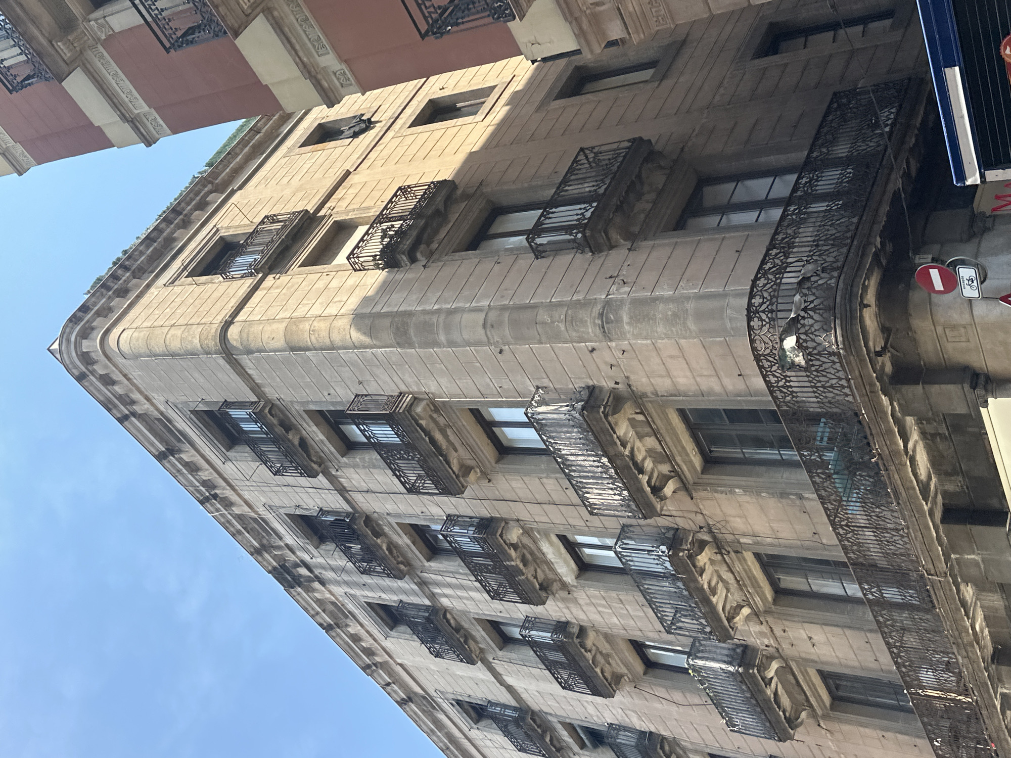 A low-angle exterior shot shows a corner of an old, light-brown stone building with multiple floors and many balconies in Barcelona, Spain. The building is made of rectangular stone blocks and has decorative moldings along its edges and above the windows. Each floor has several dark-framed windows, and many of these windows have ornate, dark metal balconies. The balconies have intricate wrought-iron railings and appear to be cantilevered out from the building's facade. The building's corner is rounded, and the balconies follow this curve. The sky above is clear and blue. To the right of the featured building, a portion of another building with a reddish-pink facade is visible, also with balconies. The lower part of the image shows a street-level view with a circular red and white traffic sign indicating no entry, and a smaller sign with a bicycle symbol below it. A blue and white stripe of what appears to be a vehicle or awning is also present at the bottom. The architecture suggests an urban European setting, likely in a historic district of Barcelona.