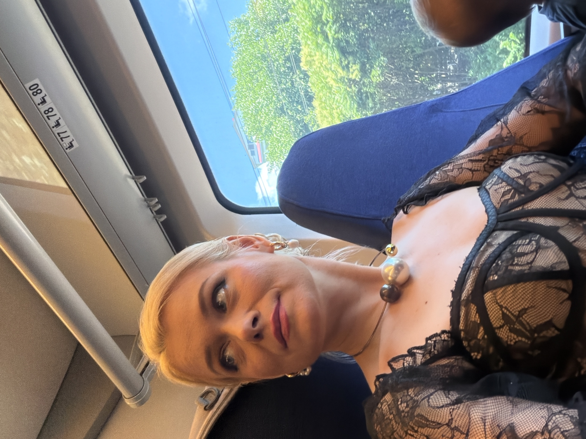 A woman with blonde hair is seated inside a train carriage, looking upwards and to her left, with a slight smile. She is wearing a black lace top with a sweetheart neckline and a statement necklace composed of large, spherical beads in shades of grey, silver, and gold. Her ears are adorned with hoop earrings with pearl-like drops. The interior of the train car is visible, with a blue seat behind her and a window to her right showcasing lush green trees and a bright blue sky. Above the window, black and white stickers indicating seat numbers are affixed to the ceiling. A portion of another person's head is visible in the lower right corner of the image, suggesting they are seated nearby. The scene is brightly lit, likely by natural daylight. The provided location of Sochi, Russia, is not discernible from the visual elements of the image.