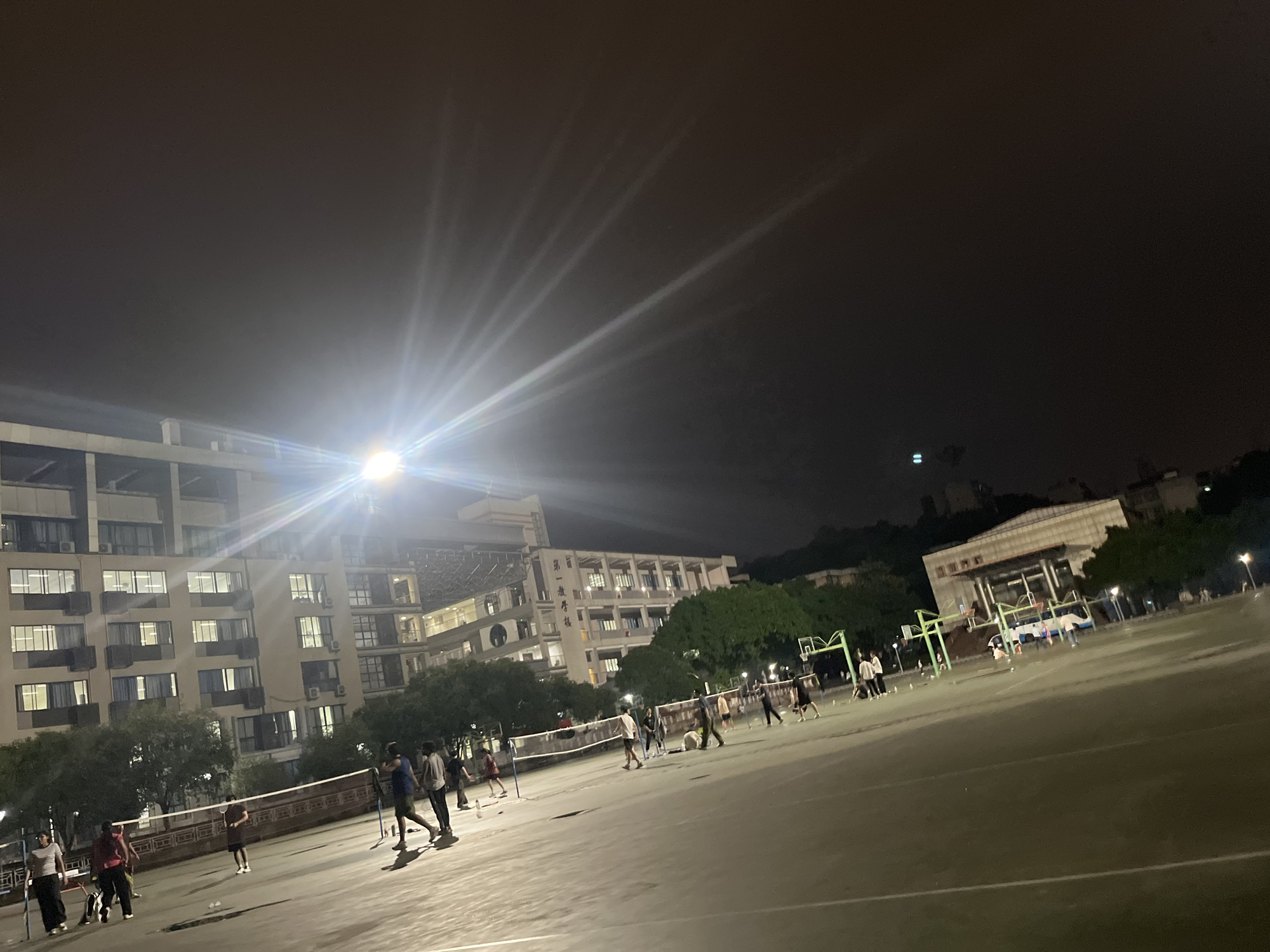 At night, a group of people are playing badminton on a court in front of buildings in Yuzhong District, China. The court is lit by overhead lights, creating strong beams of light that fan out across the dark sky. Several individuals are engaged in a badminton match, with a net stretched across the court. Other people are standing around, possibly waiting to play or observing the game. Trees are visible behind the court, and the buildings in the background are illuminated, showing multiple windows and air conditioning units. The overall scene depicts a recreational activity taking place outdoors after dark.