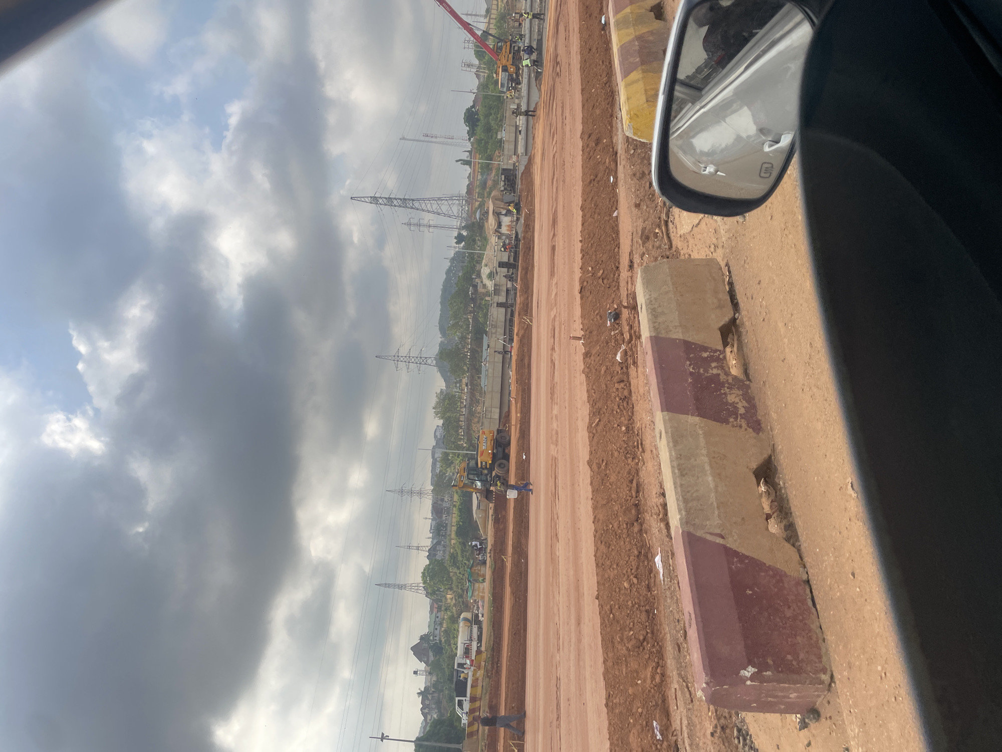 The image shows a construction site viewed from a vehicle, likely a car, in Abuja, Nigeria. The side mirror of the vehicle is visible in the upper right corner. The construction appears to be for a road, with red soil forming the base. Workers and construction vehicles, including a crane and an excavator, are present. Tall electricity pylons line the construction area. The sky is cloudy. A roadside curb with alternating red and beige paint blocks is visible on the right.
