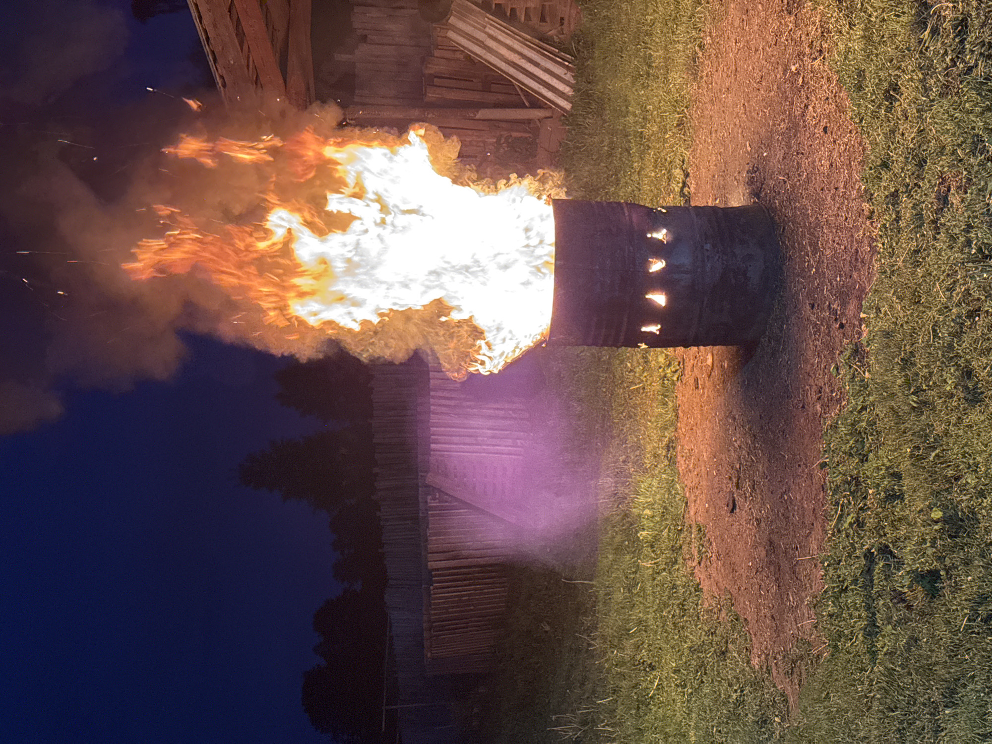 A large, intense fire blazes from a horizontally oriented, dark metal barrel in an outdoor rural setting at night or late dusk. The barrel rests on a patch of dirt and sparse grass, which transitions into lush green grass. Bright orange and white flames, accompanied by substantial grey smoke, forcefully exit the left end of the barrel, casting a strong light on the surroundings. Small, glowing embers and sparks are visible within the smoke plume.
On the visible side of the barrel, several irregular cut-out shapes allow the internal glow of the fire to shine through. These shapes are abstract, resembling elongated triangles or rudimentary symbols. A dark wooden structure, possibly a building or stacked timber, is partially visible in the background to the left, faintly illuminated by the fire. The upper left portion of the image shows a dark, deep blue night sky. The scene captures a powerful outdoor burn, potentially for disposal or heat in a location like Rusalevka, Russia. No people are visible.