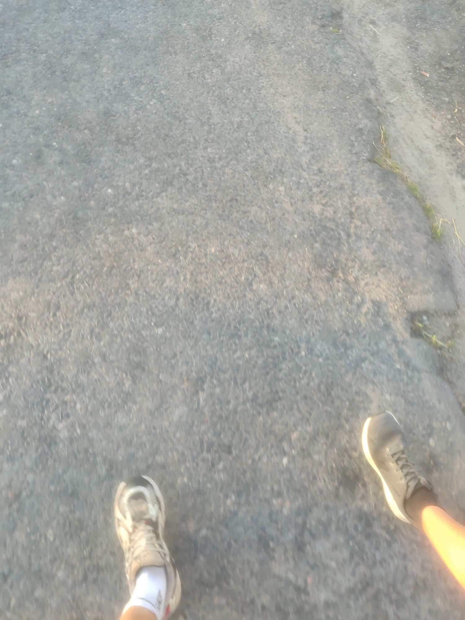 The image captures a first-person perspective of someone walking on a gray asphalt surface. The focus is on the ground and the lower legs and feet of the individual. On the left side of the frame, a white and gray athletic shoe with white socks is visible. On the right side, a dark gray athletic shoe with a white sole is visible, with a bright yellow-orange leg or light source casting a glow on it. The asphalt appears somewhat worn and textured, with some small patches of grass or debris scattered on it, particularly towards the top right of the image. The lighting suggests it is either early morning or late afternoon, with long shadows potentially present, though not clearly defined in this particular view. The overall impression is of movement or activity taking place outdoors on a paved surface.