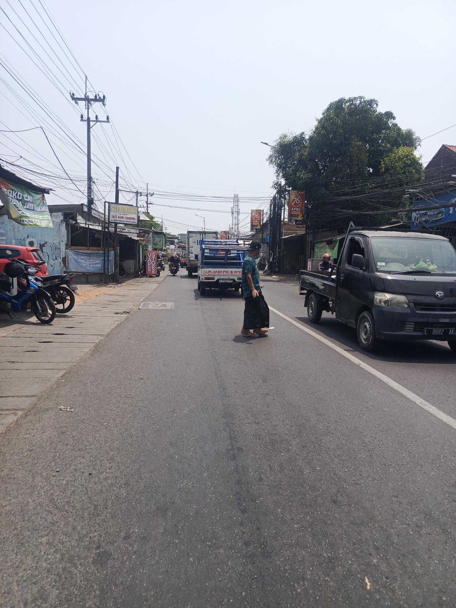 A street scene in Semampir, Indonesia, on a bright, hazy day, features a paved road with a white line marking the lane. A person dressed in a patterned shirt and sarong is crossing the road from left to right. To the right of the pedestrian, a black Daihatsu pickup truck with license plate L 3682 AK is stopped in the lane. The truck has a passenger in the cabin. In the distance, traffic is moving along the road. On the left side of the road, a sidewalk runs alongside several buildings. Motorcycles are parked on the sidewalk, including a red and black motorcycle and a blue and black motorcycle. Signs for businesses such as "QKD CAT" and "SOTO DAGING" are visible on the buildings. Overhead, numerous electrical wires are strung between utility poles. Trees are present on the right side of the scene.