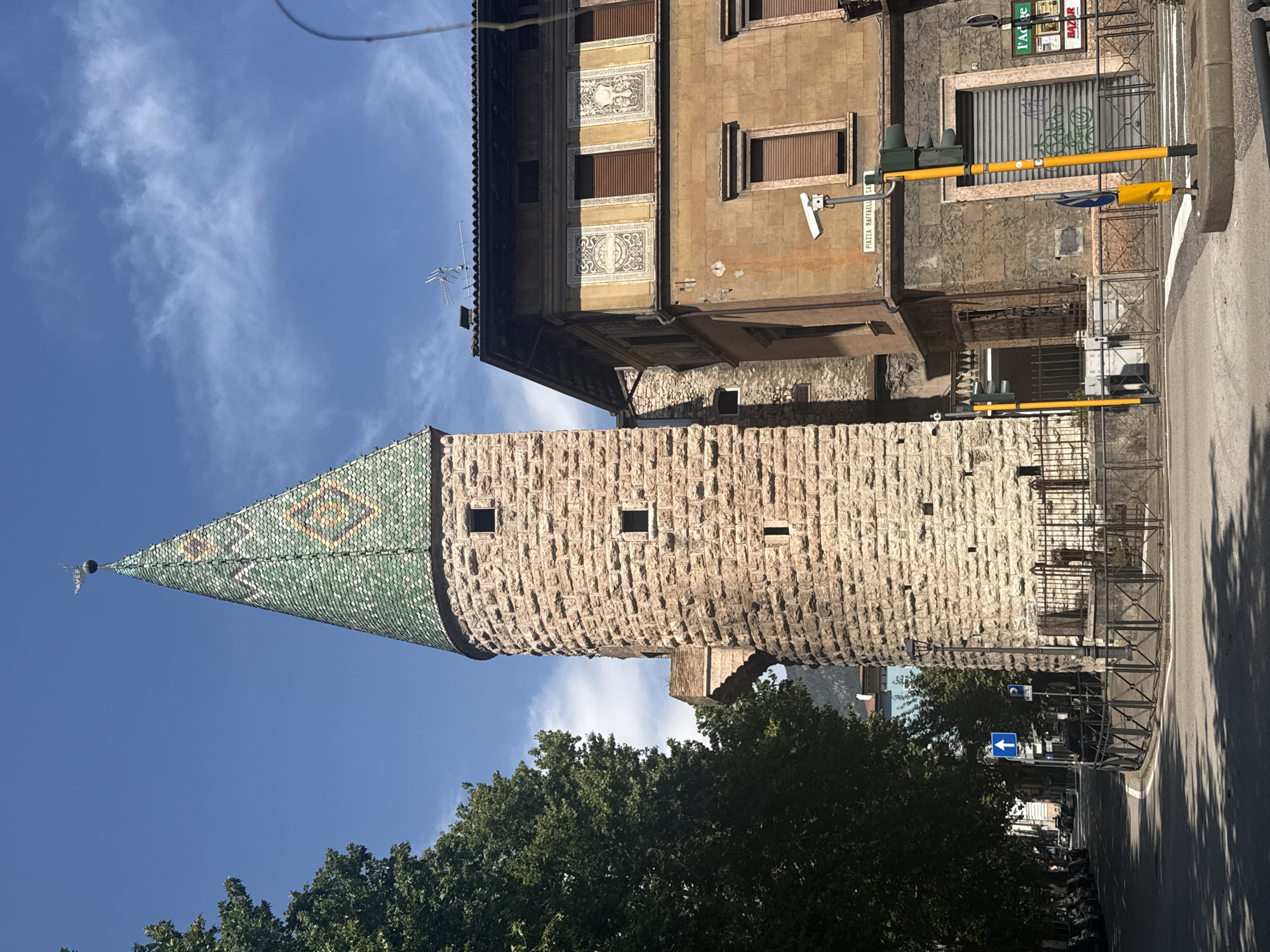The image depicts a street view in Trento, Italy, focusing on a cylindrical stone tower with a green, diamond-patterned tiled conical roof, topped by a weather vane. To the right of the tower is a light brown, multi-story building with decorative elements above the windows and closed shutters. A traffic light with yellow poles stands in the foreground. The street is paved, with a sidewalk and parked cars visible on the left side. Trees line the street, casting shadows on the road. The sky is blue with scattered white clouds. A sign on the building reads "PIAZZA RAFFAELLI SA".