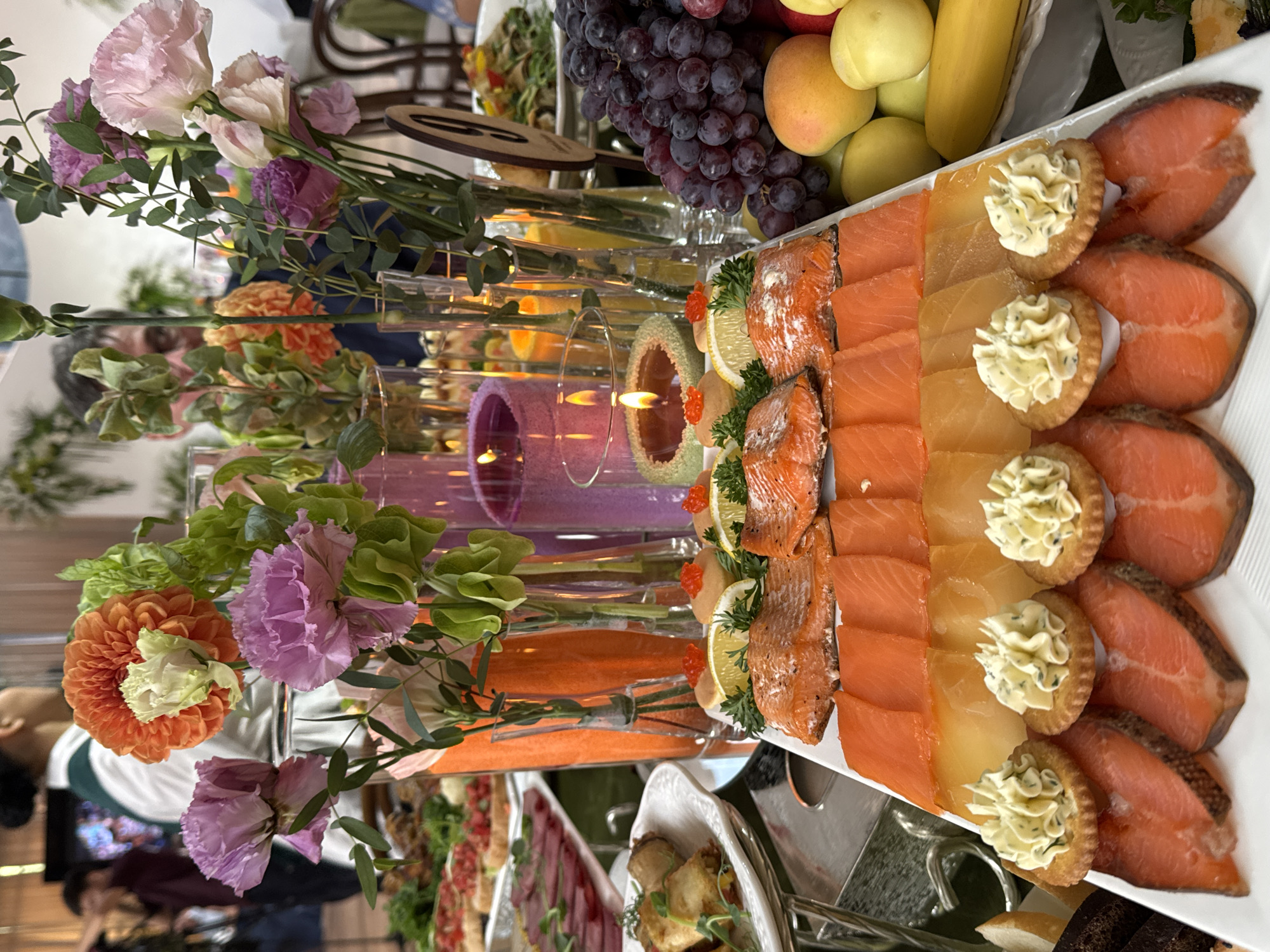 This is a photograph of a banquet table set up for a formal event, possibly a wedding or a special celebration, in Arashan, Kyrgyzstan. The table is laden with an array of food items, including a generous spread of smoked salmon, various types of appetizers, fruits, and some decorative elements.
In the foreground, a large white platter showcases meticulously arranged slices of smoked salmon. Above the salmon, there are rows of what appear to be smoked cheese slices, and on top of that, individual crackers topped with dollops of a creamy, herb-infused spread. The presentation is artful, with each cracker and topping placed with precision.
The background of the image is populated with vibrant floral arrangements in clear vases, adding a touch of elegance and color to the scene. Pink, purple, and orange flowers, interspersed with green foliage, create a visually appealing display. There are also several tall, thin vases containing what looks like liquid, possibly water or a decorative filler, and some candles with lit flames, one of which is in a green scalloped holder and topped with red caviar.
Beyond the immediate food display, other elements of the banquet are visible. To the left, there are trays with more appetizers, including what looks like rolled spring rolls and thin slices of cured meat. To the right, a bunch of dark grapes, several yellow apples, and a banana are arranged, adding a fresh fruit component to the spread. In the further background, blurry figures of people can be seen, suggesting that guests are present at the event. The overall ambiance appears to be sophisticated and festive, with a focus on a luxurious food presentation.