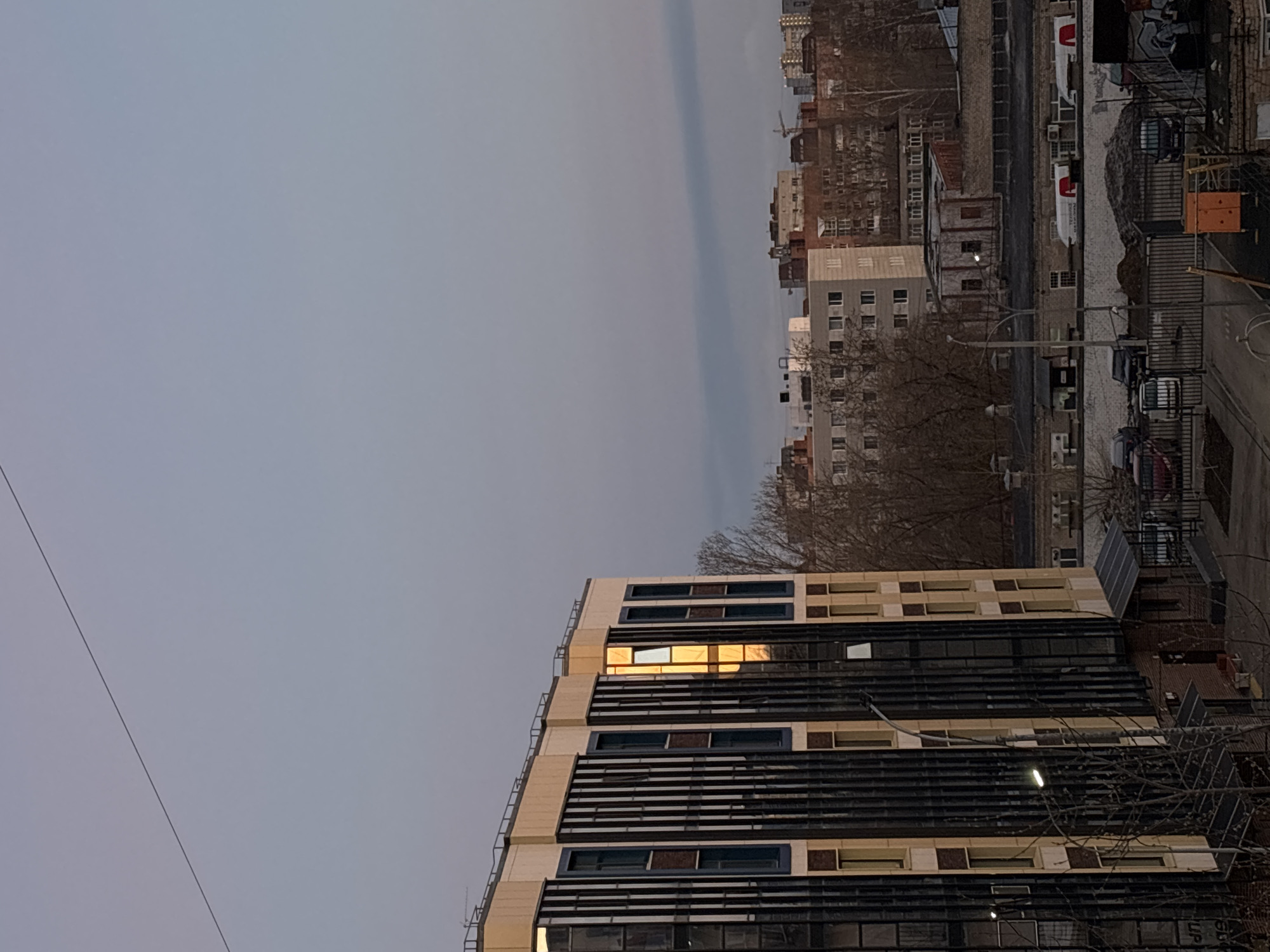 The image shows a cityscape of Perm, Russia, under a grey sky. A modern building with horizontal sunshades dominates the foreground, its windows reflecting a golden light. In the background, older buildings of varying heights are visible, interspersed with bare trees. A road with parked vehicles runs along the side of the buildings in the background. The city appears to be in a residential or mixed-use area.