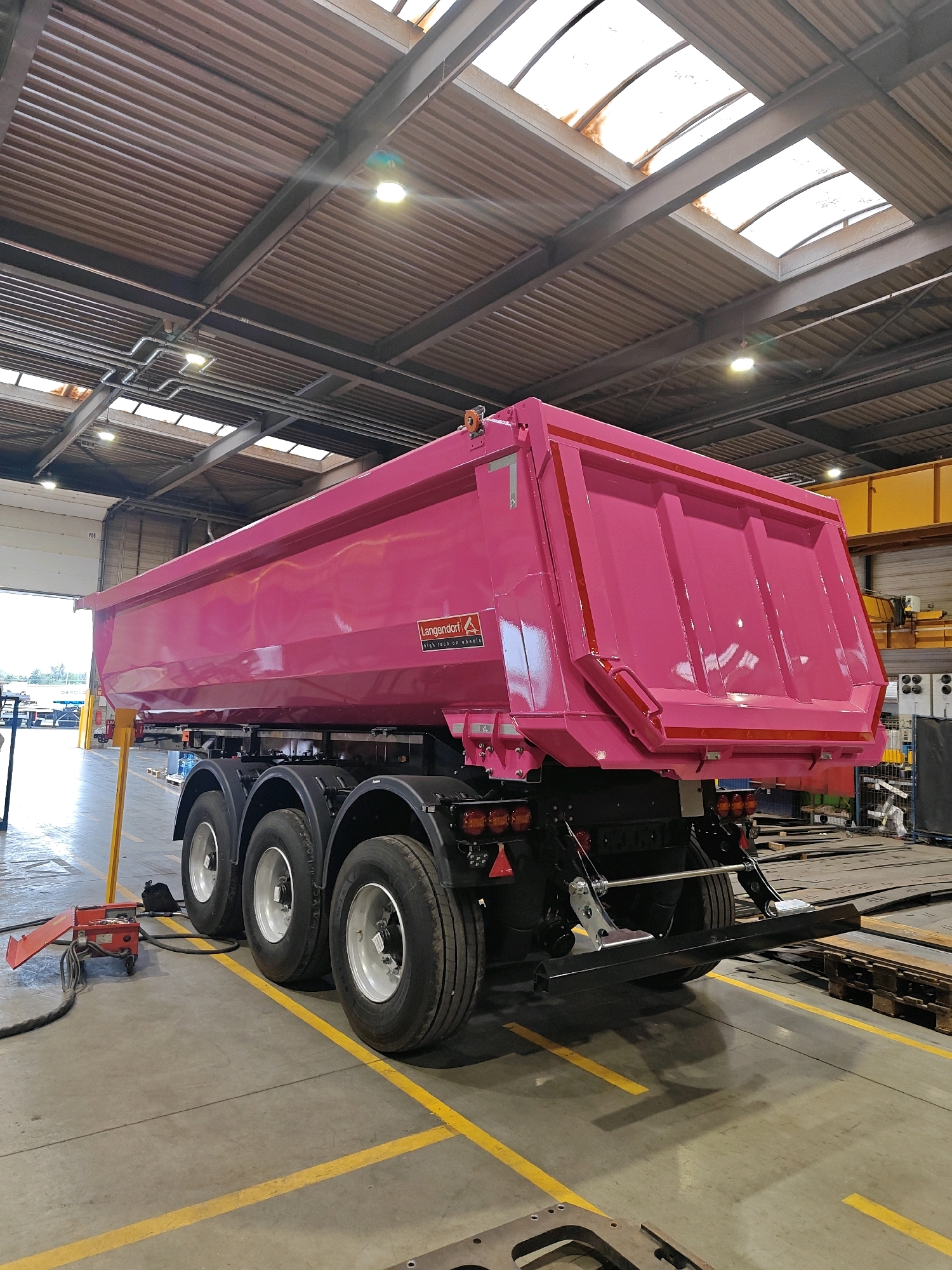 An interior view of a factory or workshop in Wieluń, Poland, shows a large, bright pink dump truck trailer. The trailer is the primary subject, positioned diagonally from the lower left to the upper right of the frame. It features multiple wheels and a large, open-top cargo bed with reinforced sides and a tailgate. The trailer displays branding with a logo and text that reads "Langendorf" and "High tech on wheels".
The environment is industrial, with a high ceiling supported by exposed metal beams. The ceiling has corrugated metal sheeting and several skylights allowing natural light to filter in. Fluorescent lights are also visible, illuminating the space. The floor is concrete, marked with yellow lines that appear to be traffic or safety guides. Various industrial equipment and materials, such as metal sheets and wooden pallets, are scattered around the workshop, suggesting ongoing construction or manufacturing. A red piece of machinery with a hose is visible in the foreground, to the left of the trailer. The overall impression is a busy and functional industrial setting.