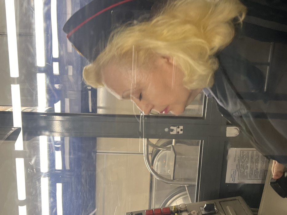 The image displays an adult female seated within a glass-enclosed operator's booth at a metro station in Saint Petersburg, Russia. The woman, shown in right profile, is middle-aged to older with bleached blonde hair. Her eyes are closed and lips slightly parted, indicating a state of rest or sleep. She is wearing a dark, possibly uniform, jacket. A dark uniform cap with a prominent red stripe is visible behind her head, resting on a surface. Her left hand is positioned near a dark control handle or joystick, which is part of a grey control panel featuring several red and white circular buttons and a toggle switch. A document with Cyrillic text is affixed to the lower portion of the booth's interior. Beyond the glass partition, a large, operational escalator with polished metal handrails and steps is visible in the background, ascending upwards. The escalator area features light-colored walls and overhead horizontal fluorescent light fixtures.