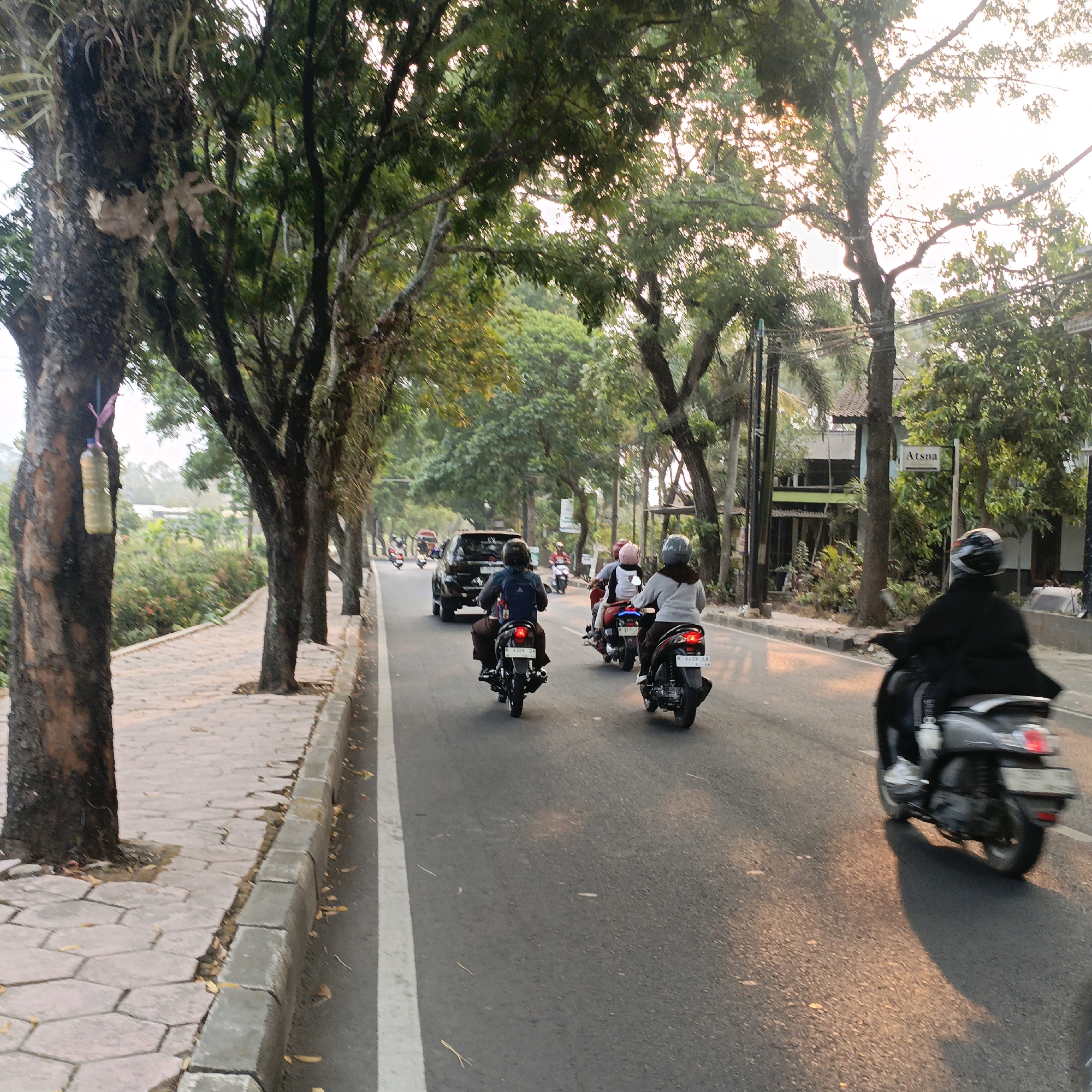 The image shows a street scene in Salatiga, Indonesia. The street is lined with trees on the left side, with a paved sidewalk. The road is asphalt and has a white line marking the lane. Several motorcycles are on the road, with riders wearing helmets. In the background, there are buildings and more trees. The sun is setting, casting a warm light on the scene. One of the trees on the left has a plastic bottle hanging from it, tied with a pink string. A sign that reads "Atsna" is visible on the right side of the street.