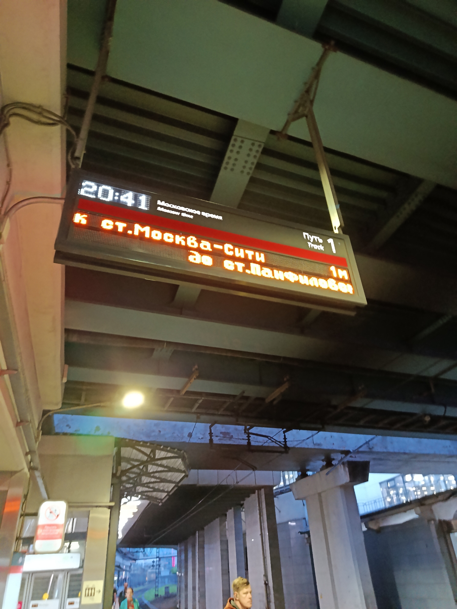 An outdoor electronic display board shows train departure information at a station. The display board shows the time as 20:41 and "Moskow time". Below that, it lists the destination as "to st. Moskva-City". Further down, it indicates the track number as "1" and "1M" which likely means 1 minute away. The station appears to be underground or covered, with concrete pillars and overhead structures visible. In the background, there are train tracks and buildings in the distance, suggesting an urban environment. Two individuals are partially visible in the lower portion of the image, appearing to be passengers at the station.