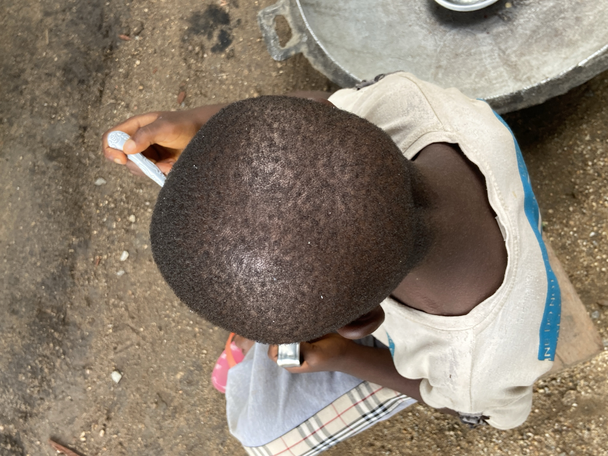 An overhead shot captures a young person seated outdoors in Mararaba, Nigeria. The primary focus is on the top of their head and upper torso. The person has very short, dark hair and dark skin. They are wearing a light-colored, possibly off-white, sleeveless top that features a prominent blue vertical stripe with fragmented, illegible text down its side. Beneath this, a grey garment with a distinct plaid pattern, reminiscent of Burberry, is visible around their lap. Pink flip-flops are worn on their feet, partially visible under the plaid garment.
In their right hand, the person holds a small, white, decorative spoon with an embossed pattern on its handle. Their left hand is also near the spoon. In the upper right portion of the frame, a large, dark metallic pot or basin with sturdy handles is present, containing a smaller, shiny metallic dish. The ground is an unpaved area of light brown and grey dirt, interspersed with small pebbles and some organic debris. The scene is brightly illuminated, indicating daytime. The activity appears to involve holding the spoon, possibly in preparation for or during a meal, though no food is directly visible on the spoon.