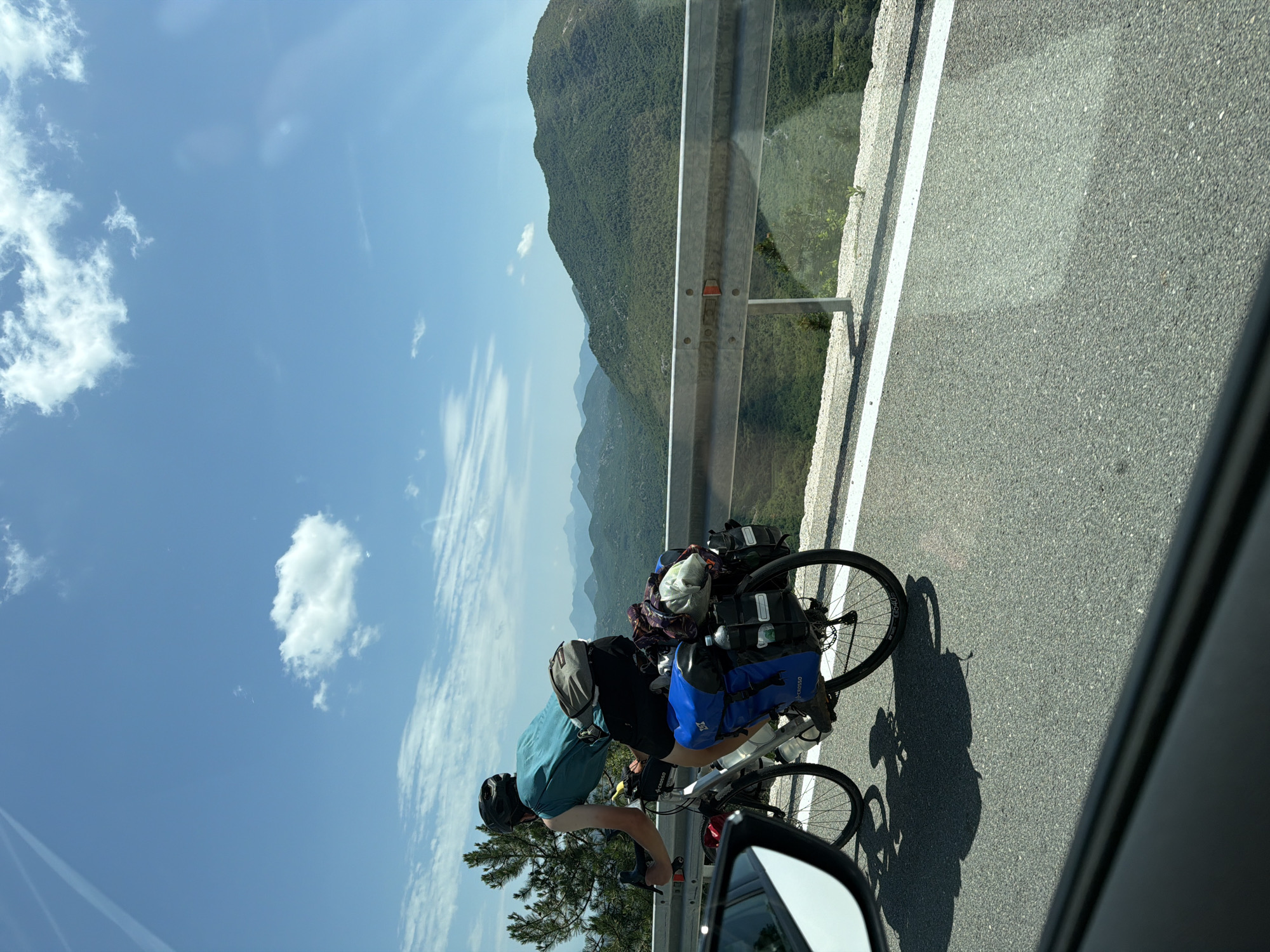 A person on a bicycle with panniers rides along a road on a sunny day in Dobrsko selo, Montenegro. The cyclist, wearing a helmet and a teal shirt, is seen from behind and to the side. The bicycle is equipped with multiple bags, indicating a long journey. In the background, rolling green hills and mountains are visible under a blue sky with scattered clouds. A metal guardrail separates the road from the scenic vista. The image is captured from a moving vehicle, as evidenced by the side-view mirror and the foreground framing.