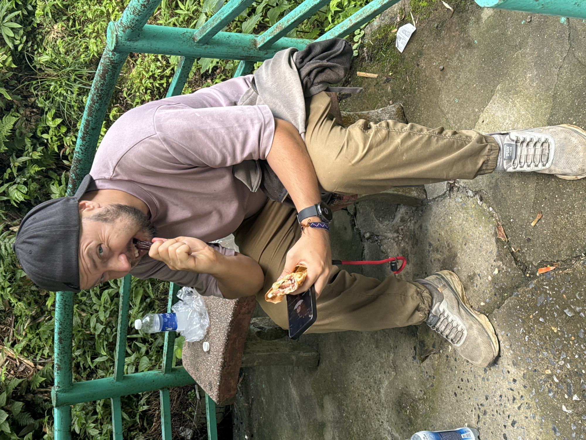A man sits on a stone bench next to a green railing in what appears to be a semi-natural setting, potentially in Sa Pa Ward, Vietnam. He is wearing a faded purple long-sleeved shirt, tan pants, grey sneakers, and a black baseball cap. He is holding a phone and eating a wrapped snack, while a water bottle sits on the bench next to him. A grey hooded sweatshirt is draped nearby. The surrounding environment includes dense greenery and a concrete area.
